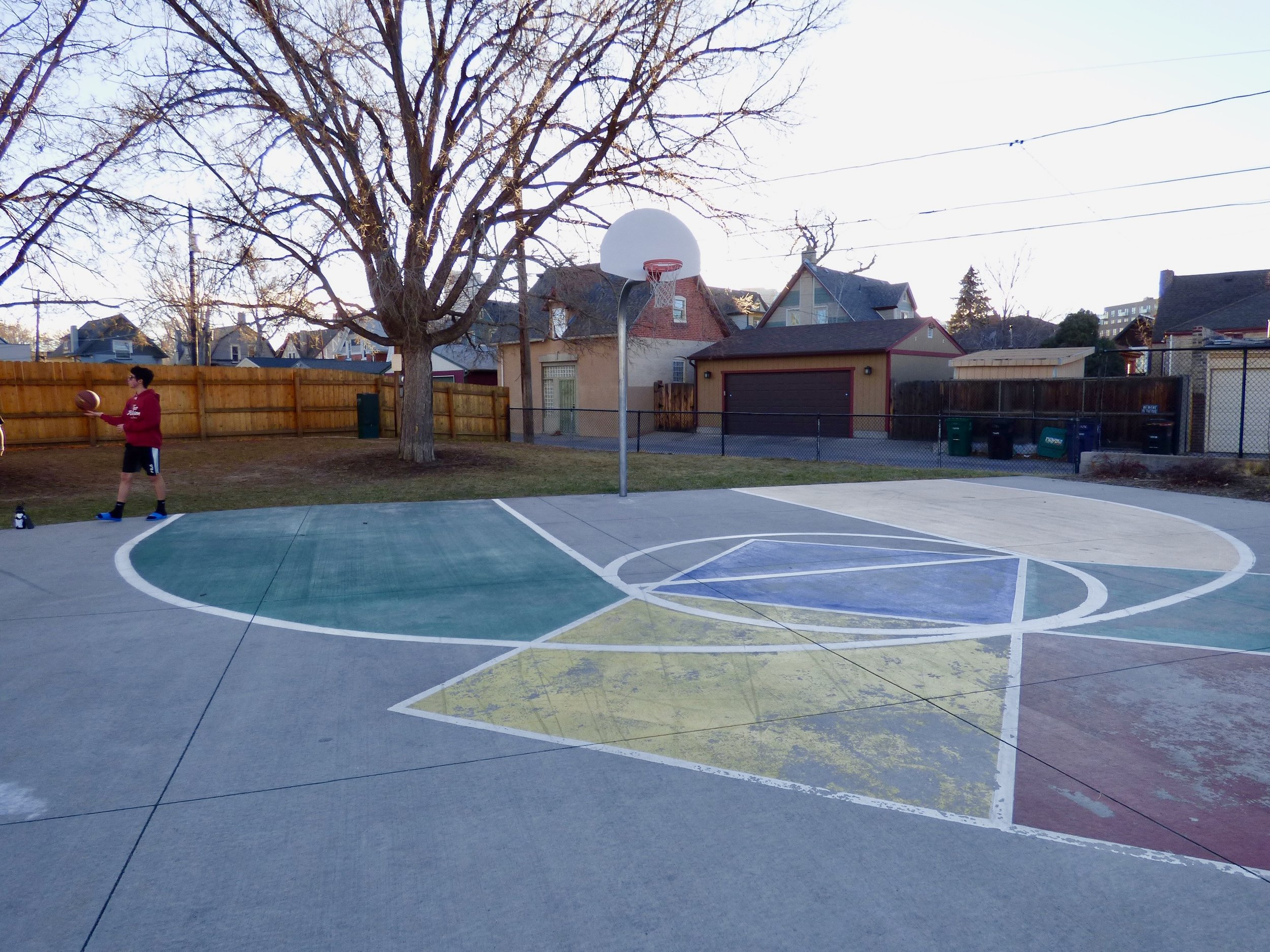 An outdoor basketball court with a young boy holding a basketball, a large tree, residential houses, and trash bins in the background.