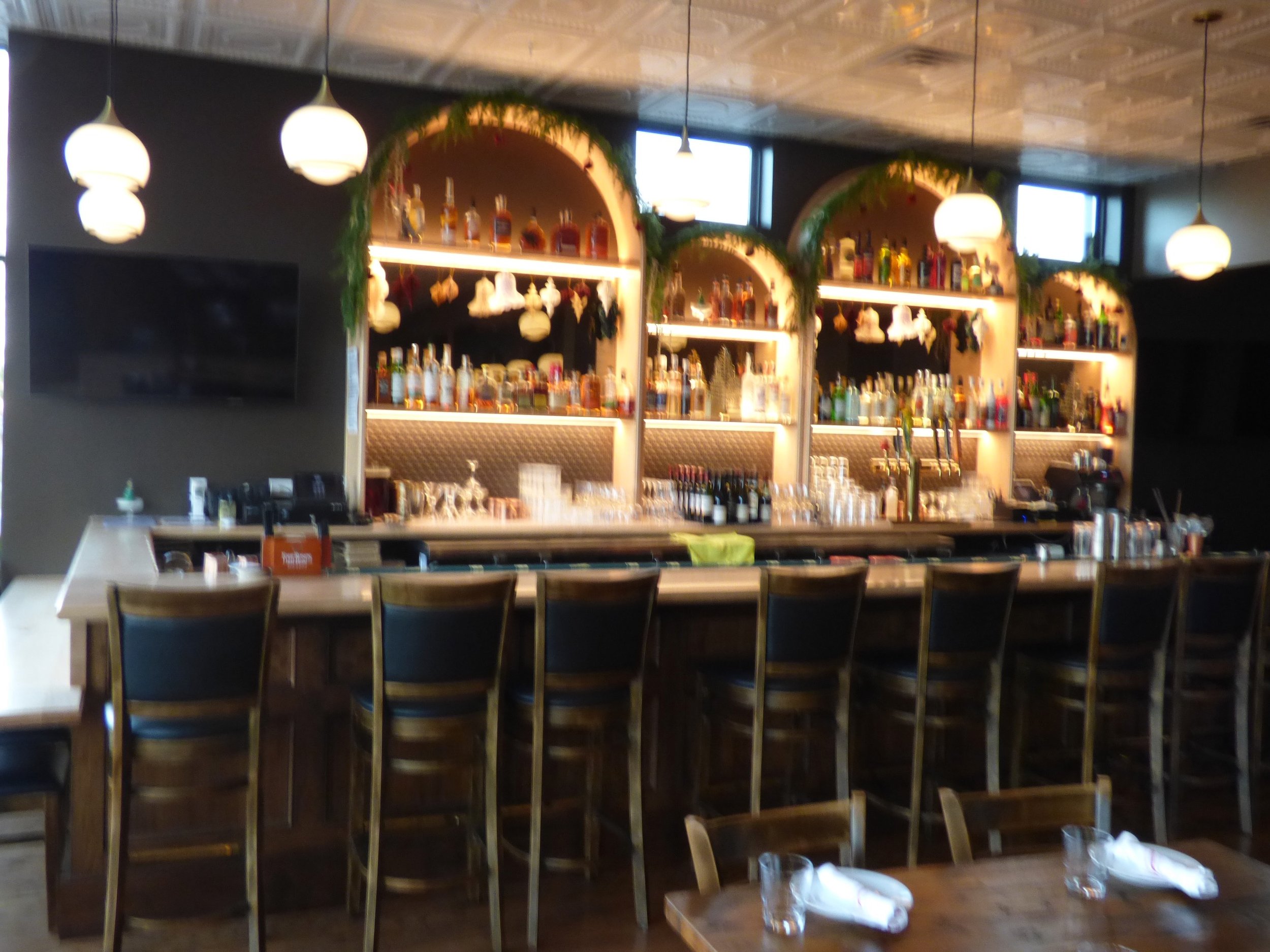 A bar with a wooden counter and high chairs, behind the bar are illuminated shelves filled with various bottles of alcohol, decorated with green garlands and holiday ornaments at Whit's End, Denver, CO 80205.