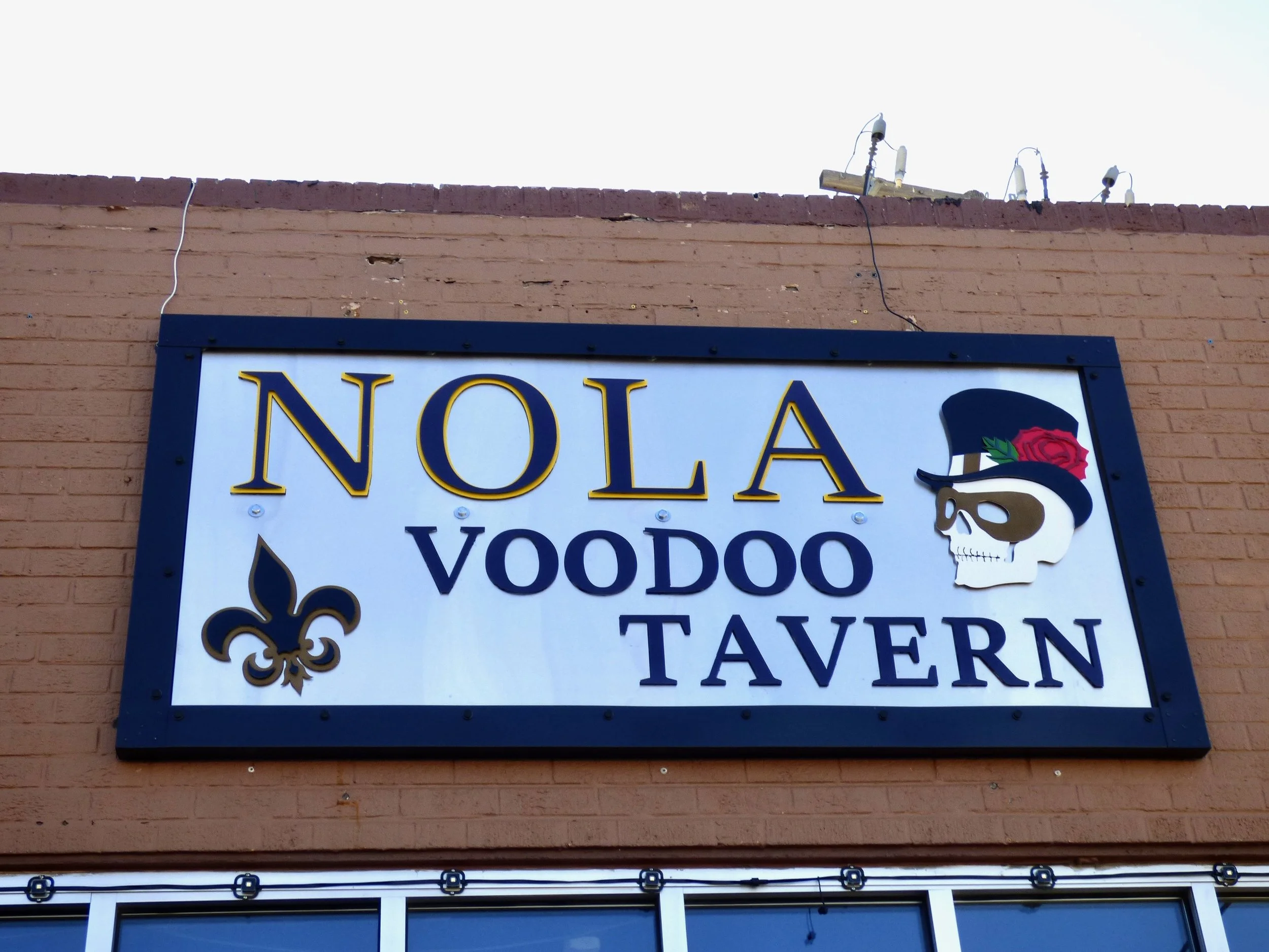 Sign for NOLA Voodoo Tavern with a skull wearing a black hat with a red rose and green leaves, and a gold fleur-de-lis symbol on a brick wall at NOLA Voodoo Tavern, Denver, CO 80205.