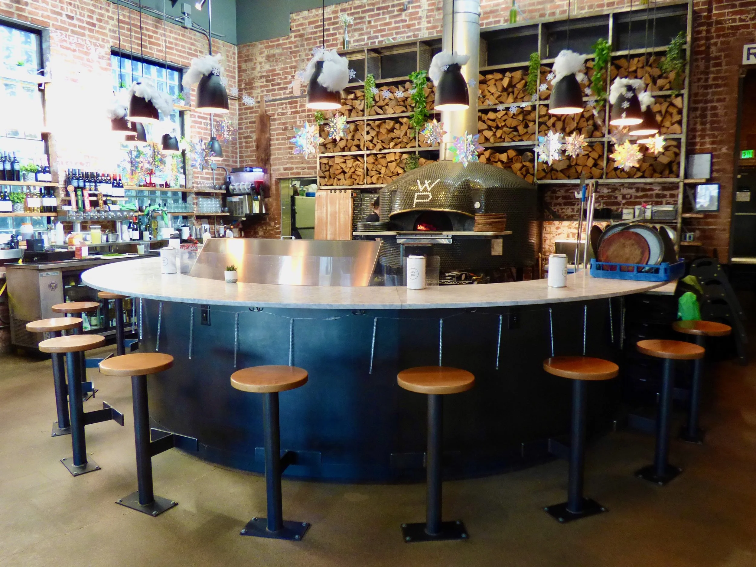 Interior of a modern restaurant with a round bar counter, wood seating stools, brick walls, shelves with wine bottles, a pizza oven, and decorative snowflake and holiday ornaments.
