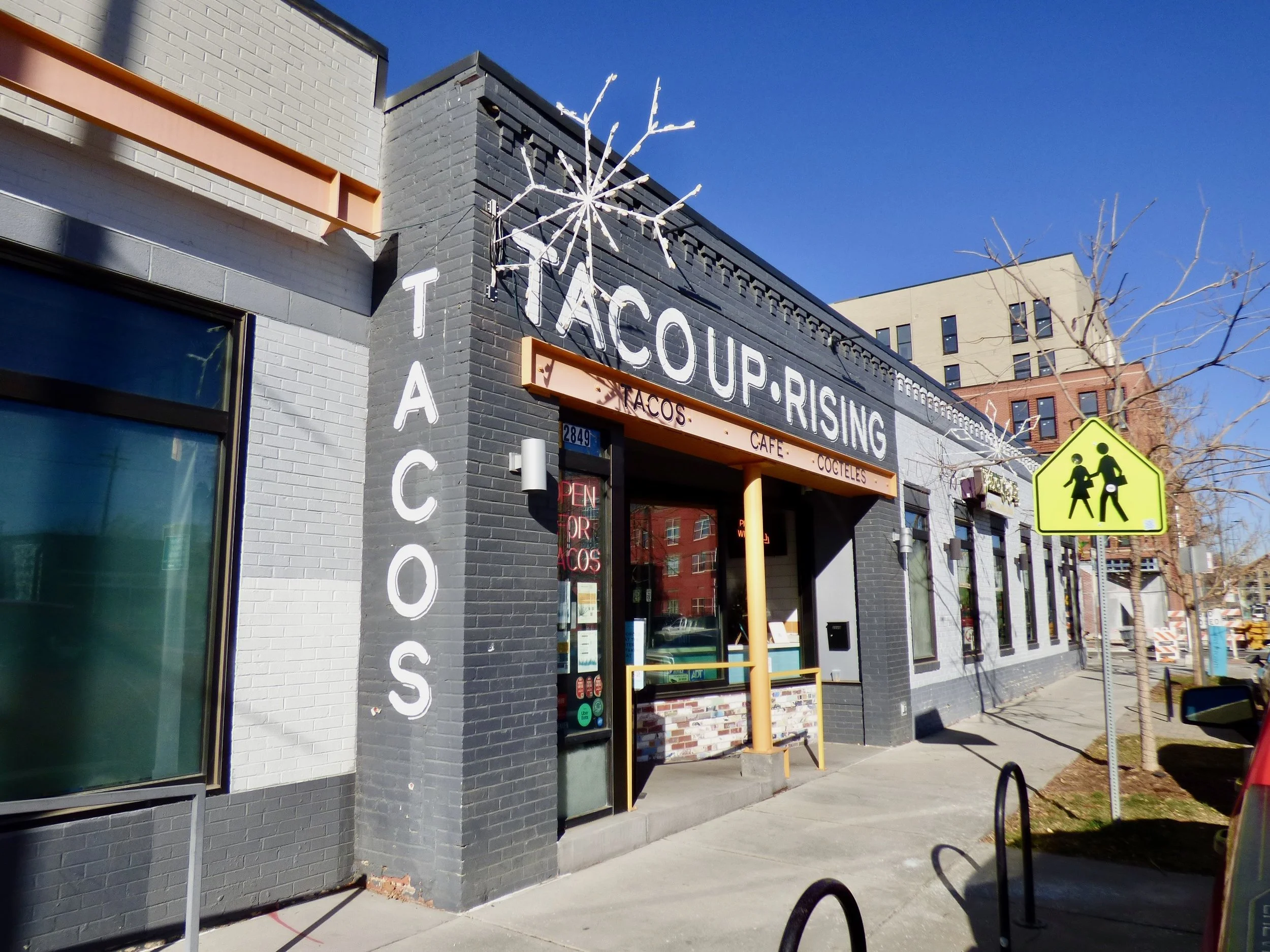 Exterior of a restaurant called Tacoup Rising with a black and white brick facade, a yellow pole at the entrance, and a yellow pedestrian crossing sign on the sidewalk. The restaurant sign indicates it serves tacos, cafe, and cocktails.
