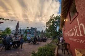 A street scene with outdoor seating, people dining, and a red building with the word 'Gotham' on it, under a partly cloudy sky at The Plimoth, Denver, CO 80205.