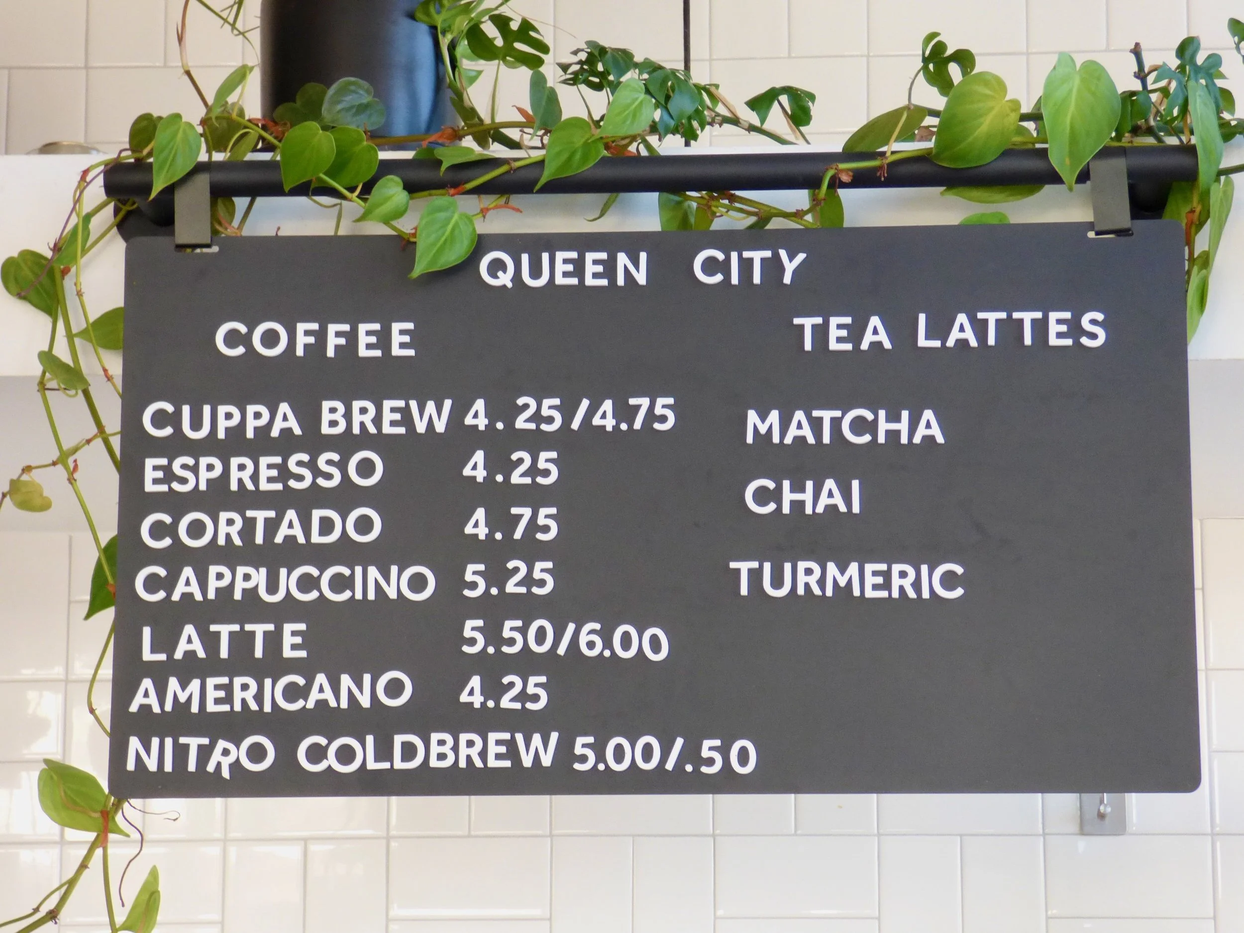 Black menu board with white text listing coffee and tea latte options, prices, and flavors at a café, decorated with green leaves hanging above at Queen City Collective Coffee House, Denver, CO 80205.