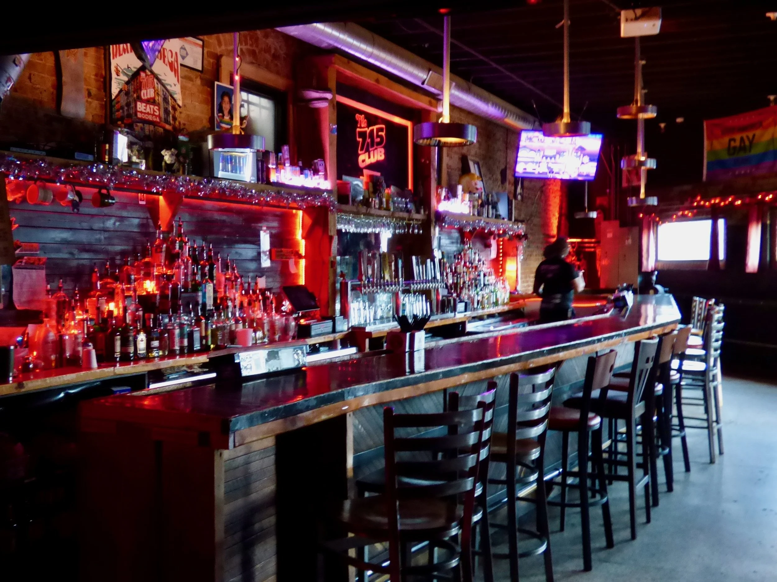 Empty bar with liquor bottles, bar stools, bartender, TV screens, and rainbow LGBTQ flag inside a nightclub or bar at 715 Club, Denver, CO 80205.