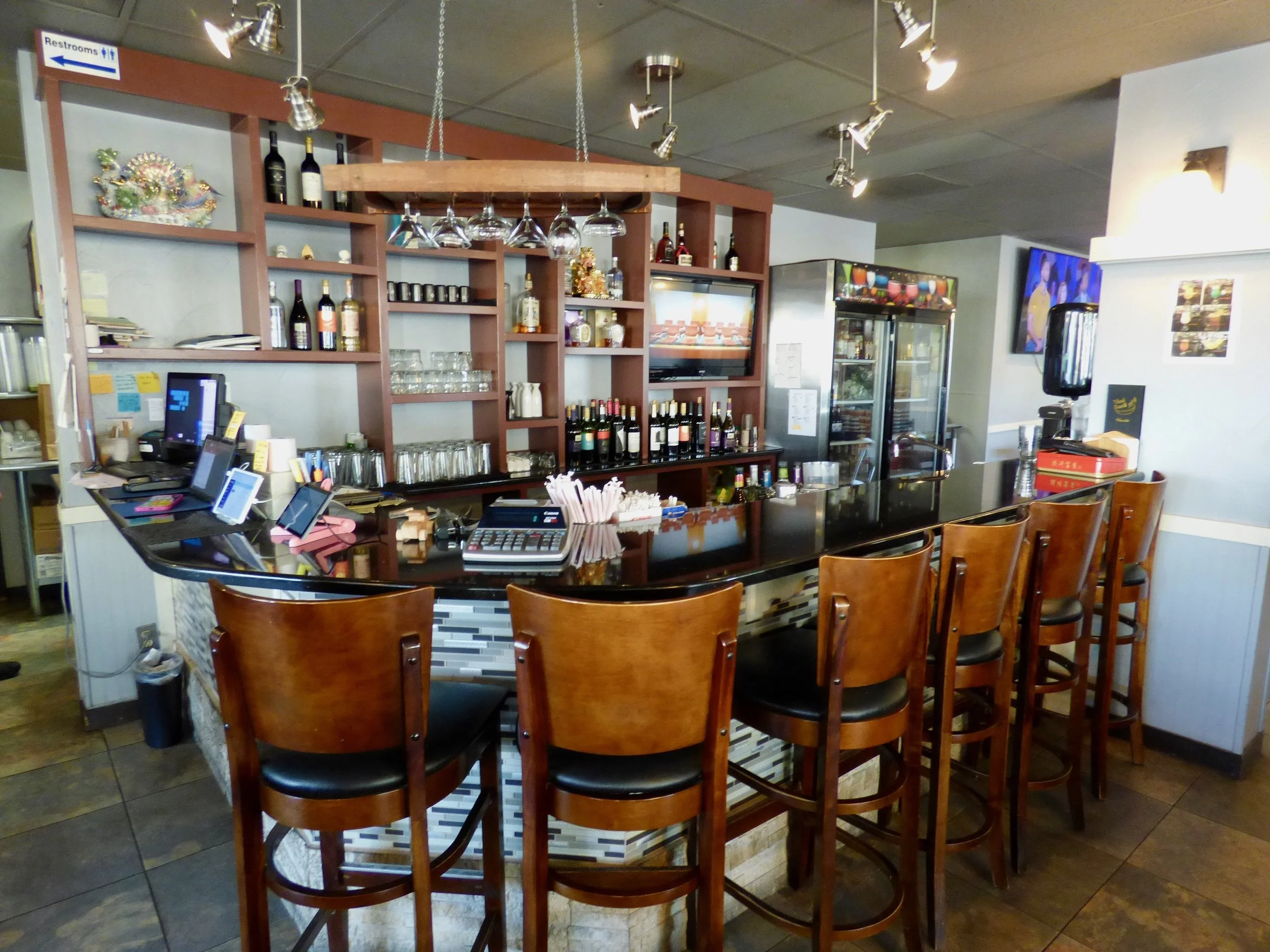 Empty bar counter with wooden chairs, liquor bottles, glasses, and a TV in a restaurant.