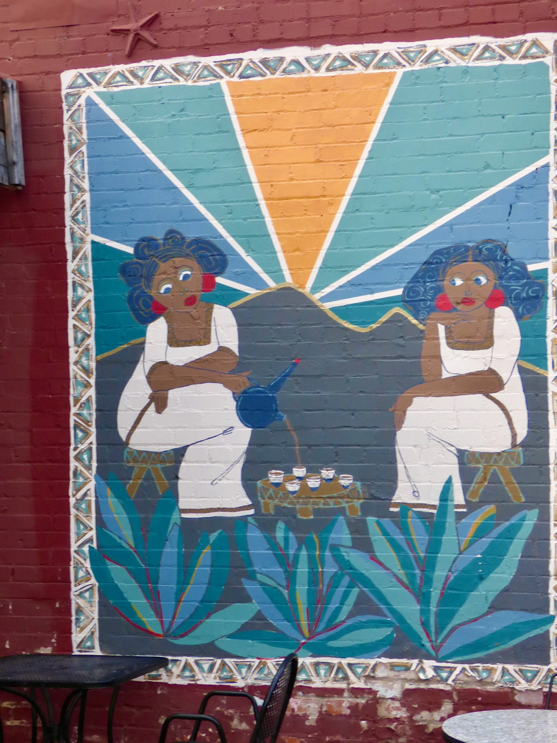 A colorful mural on a brick wall featuring two women with curly hair sitting on stools, with a mountain and sunrise or sunset in the background, surrounded by plants and decorative border at Whittier Cafe, Denver, CO 80205.