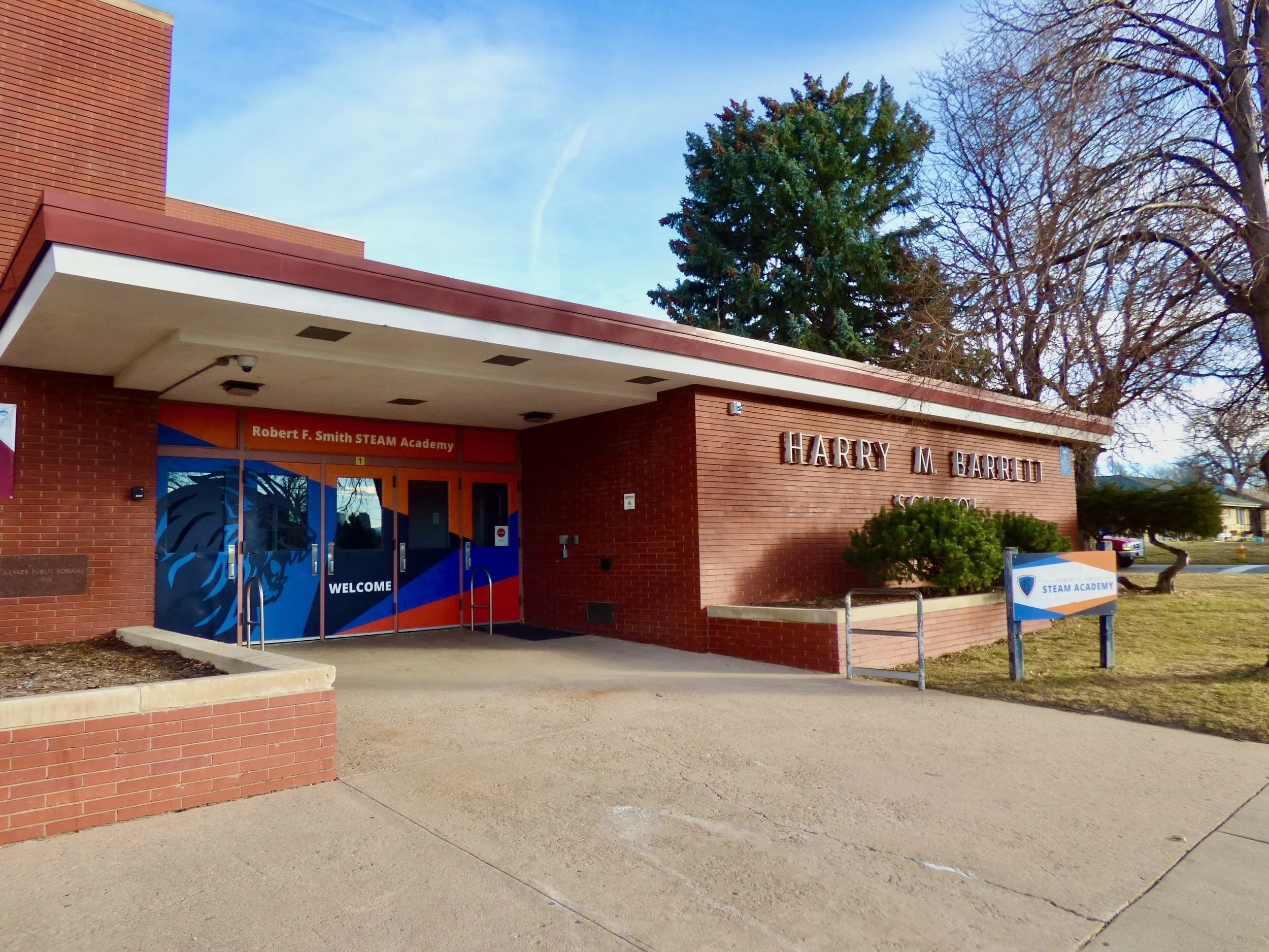The brick exterior of Harry M. Barrett School, with a colorful entrance sign for Robert F. Smith STEAM Academy, and a small garden bed with a shrub in front.