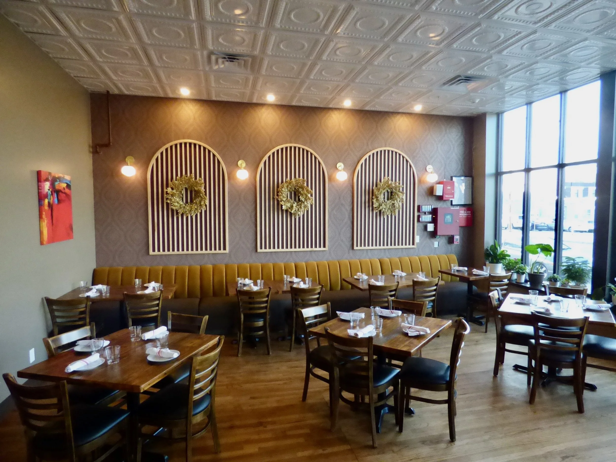 Empty restaurant dining area with wooden tables and chairs, decorated with napkins and glasses, large window with potted plants, yellow banquette seating, festive wreaths on the wall, and subtle wall lighting at Whit's End, Denver, CO 80205.