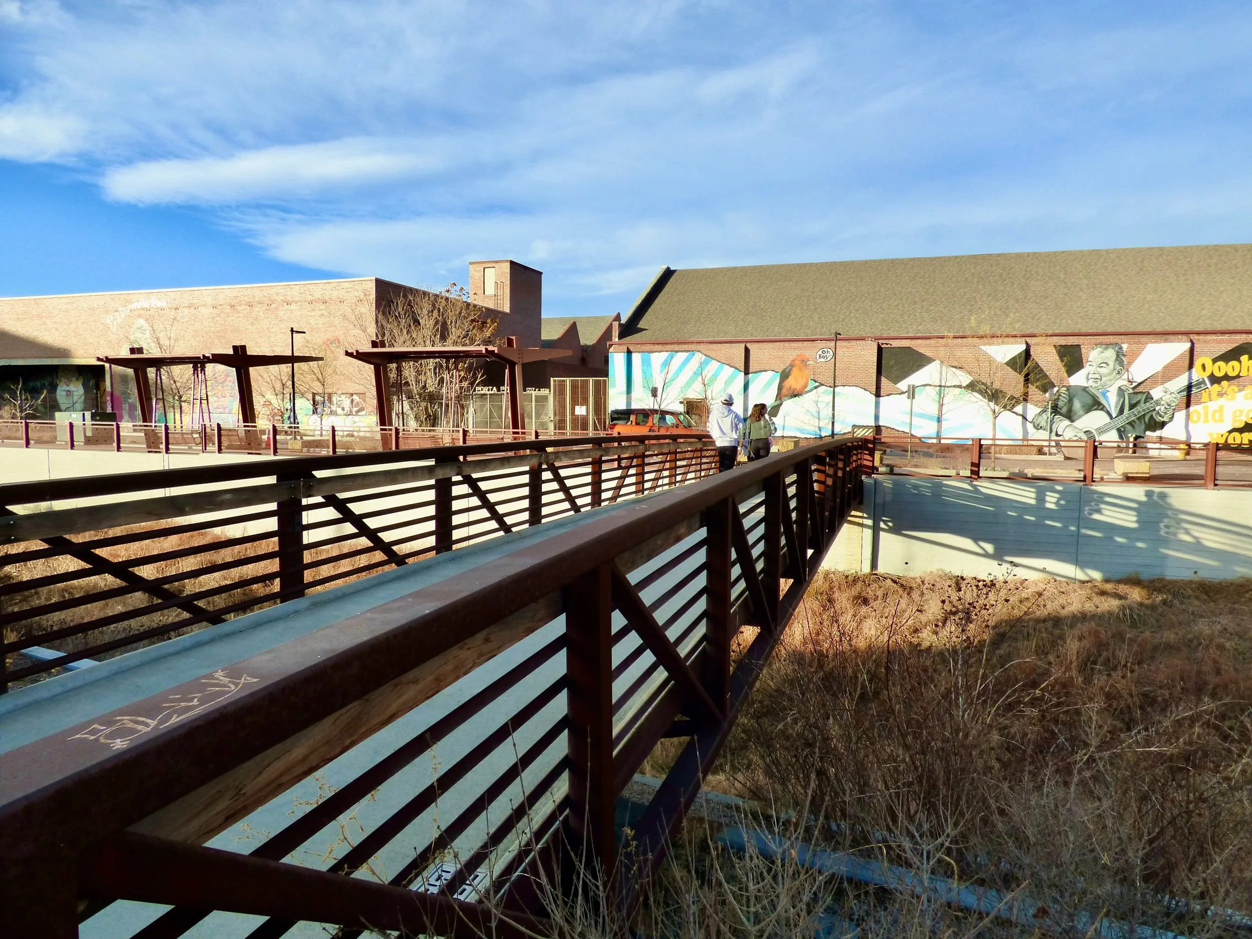 An outdoor scene featuring a wooden pedestrian bridge, some people walking, a mural on a brick building wall, blue skies, and dry vegetation in the foreground in 39th Ave Greenway, Denver, CO 80205.