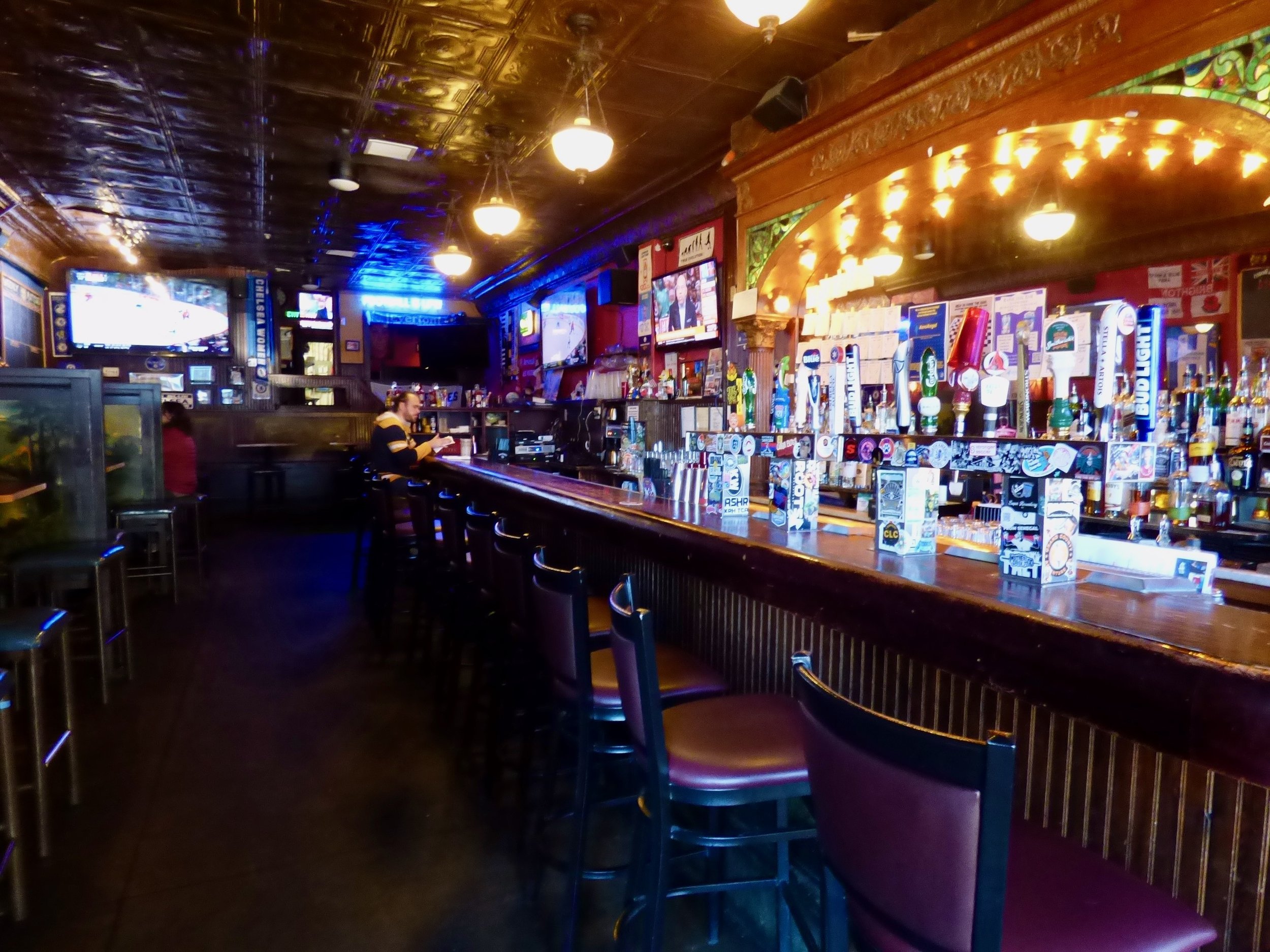 Empty bar with a single person sitting at the counter, multiple TV screens displaying sports, beer taps, dark wooden bar counter, and decorative ceiling lights in a dimly lit pub at The British Bulldog, Denver, CO 80205.