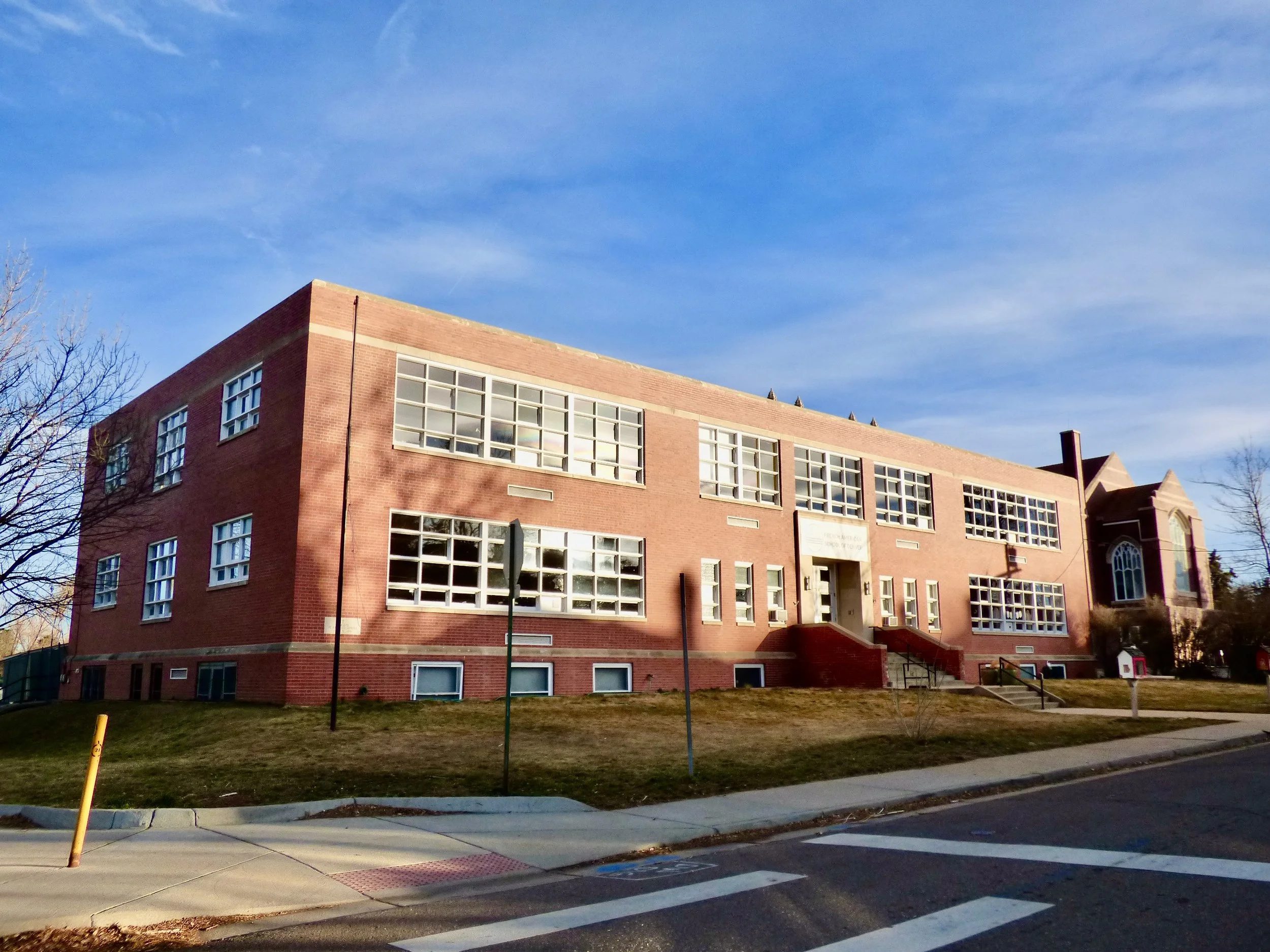 A large brick school building with multiple rectangular windows and an arched window on the right, set against a blue sky with some clouds, surrounded by grass and pavement at French American School of Denver, Denver, CO.