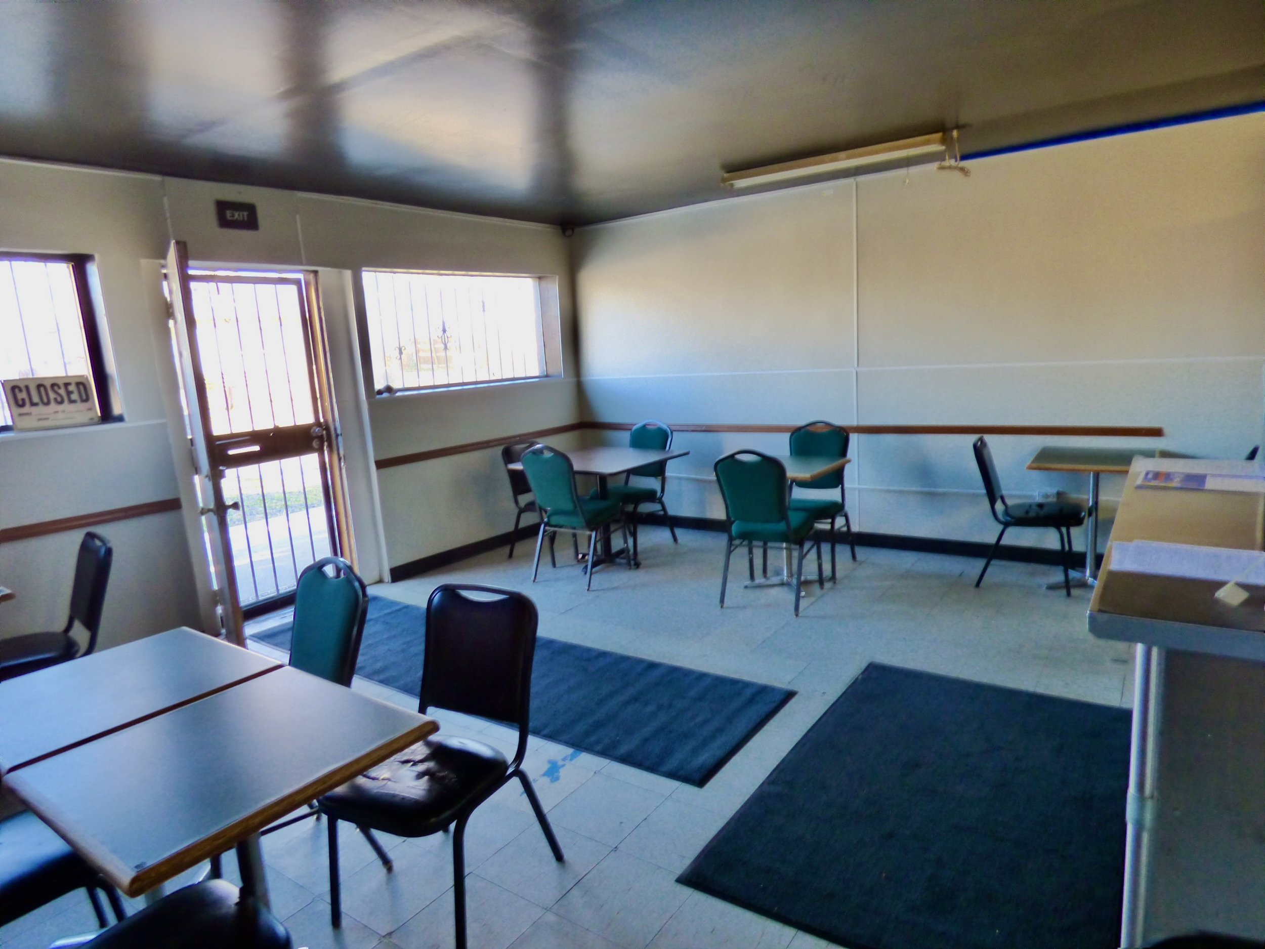 Empty dining area or lobby with tables and chairs, near entrance door with a 'Closed' sign, windows with bars, and dark mats on tiled floor.