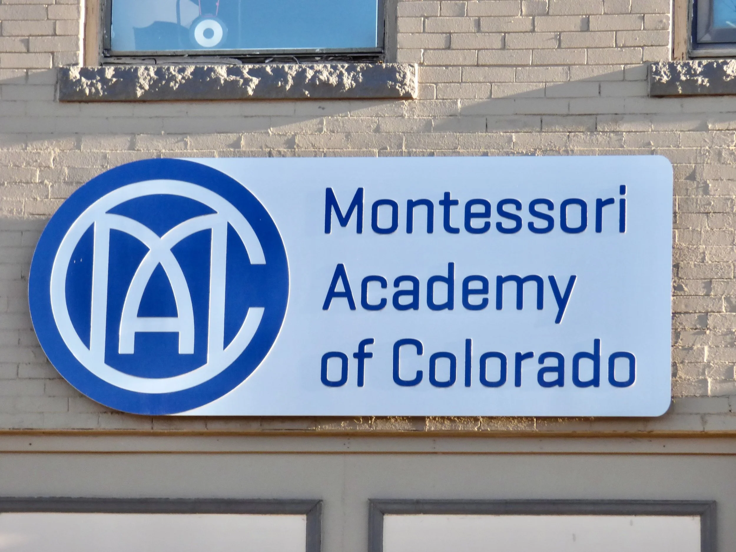 Sign for Montessori Academy of Colorado, featuring a blue circular logo with white letters 'M', 'A', and 'C' overlapping, mounted on a brick building at Denver, CO 80205.
