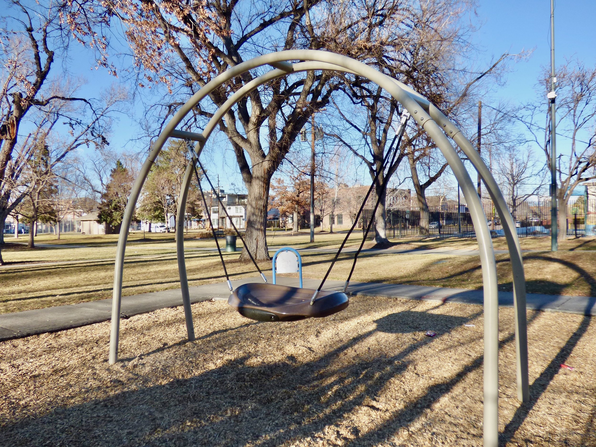 An empty playground swing set with a large tree and residential buildings in the background on a sunny day.