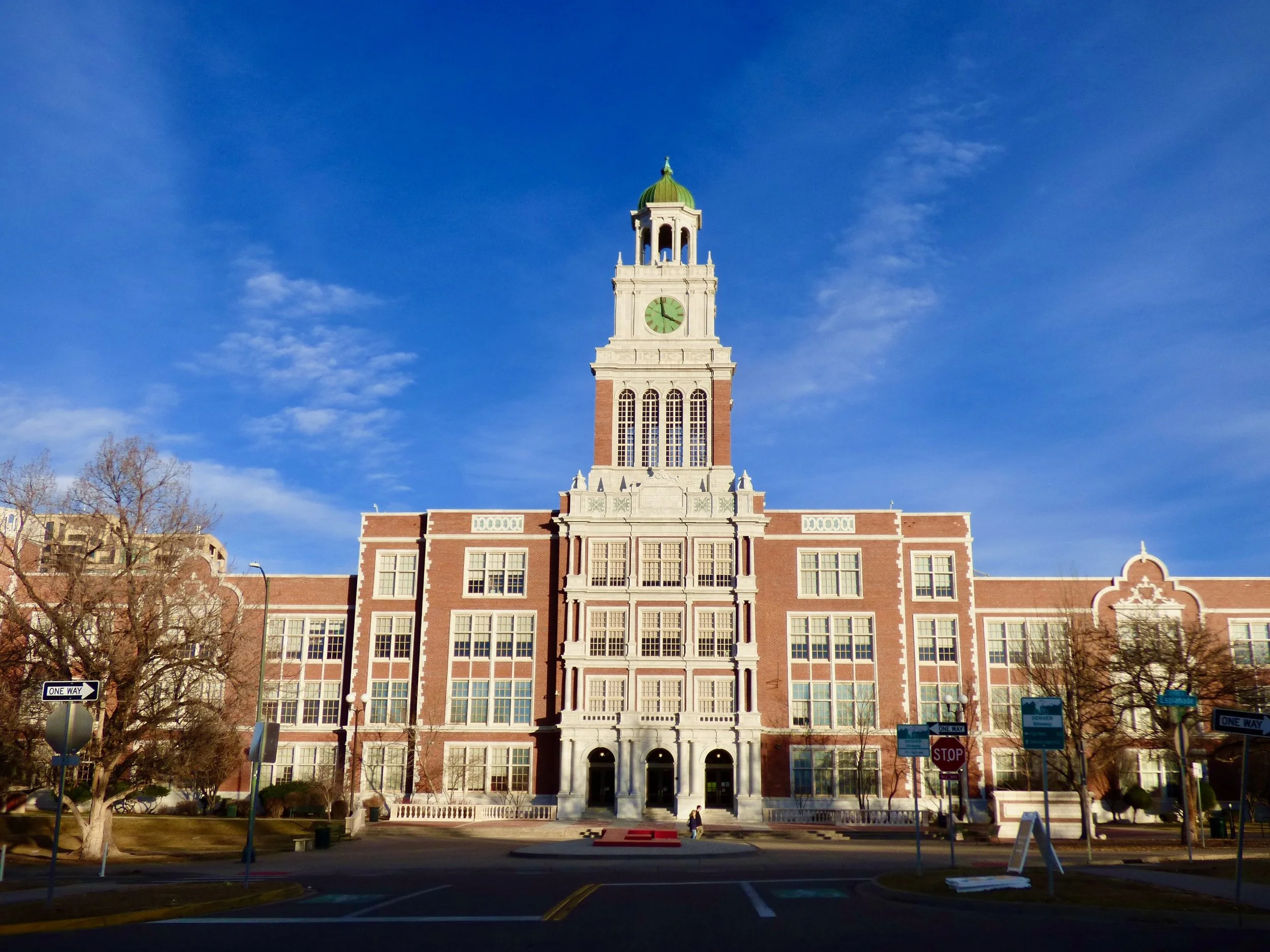 A large historic building with a clock tower under a clear blue sky, surrounded by trees and street signs at East High School, Denver, CO.