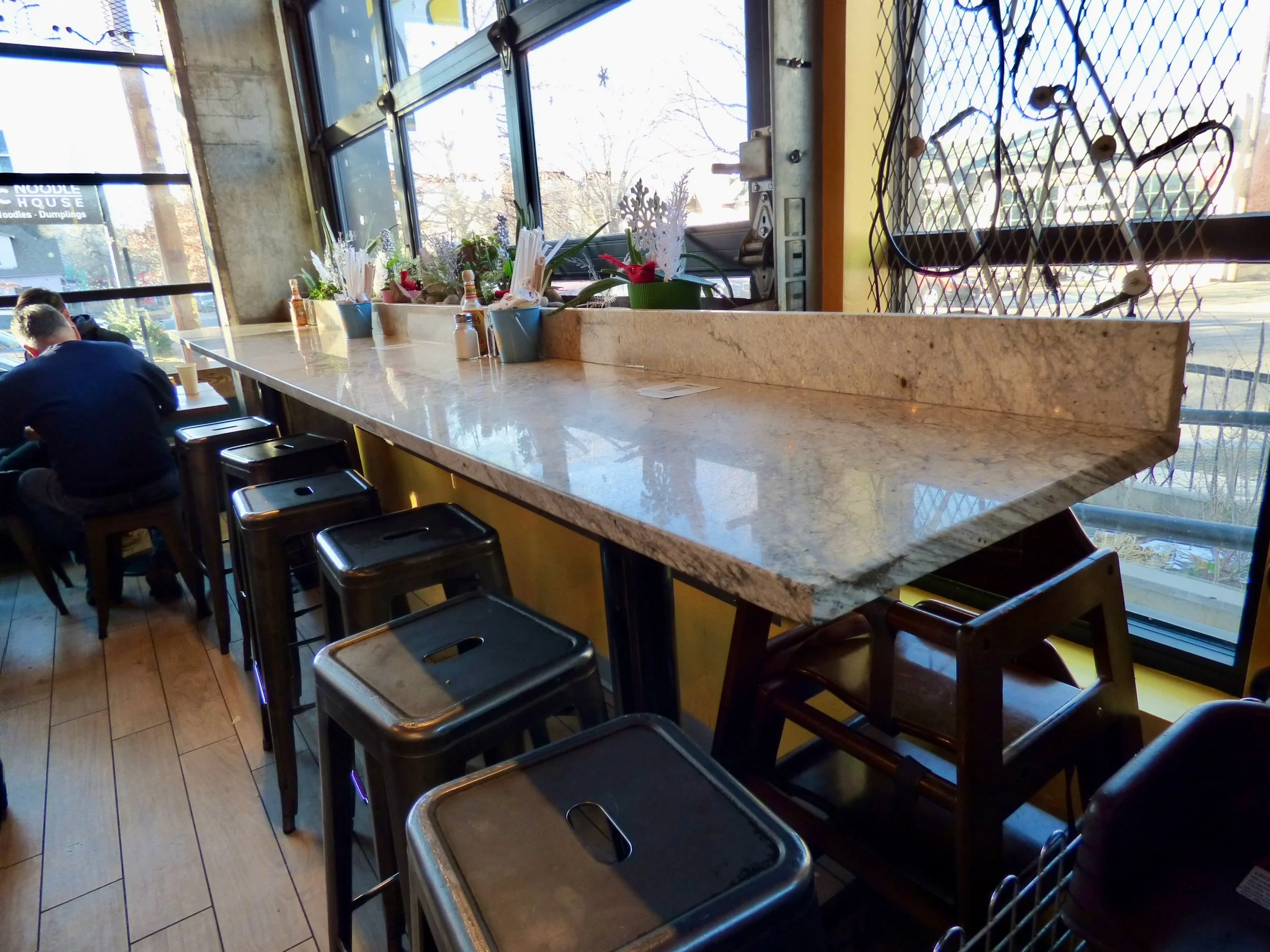 Interior of a restaurant with a long marble counter by large windows, decorated with potted plants and napkin holders, with several black stools and a few patrons seated.