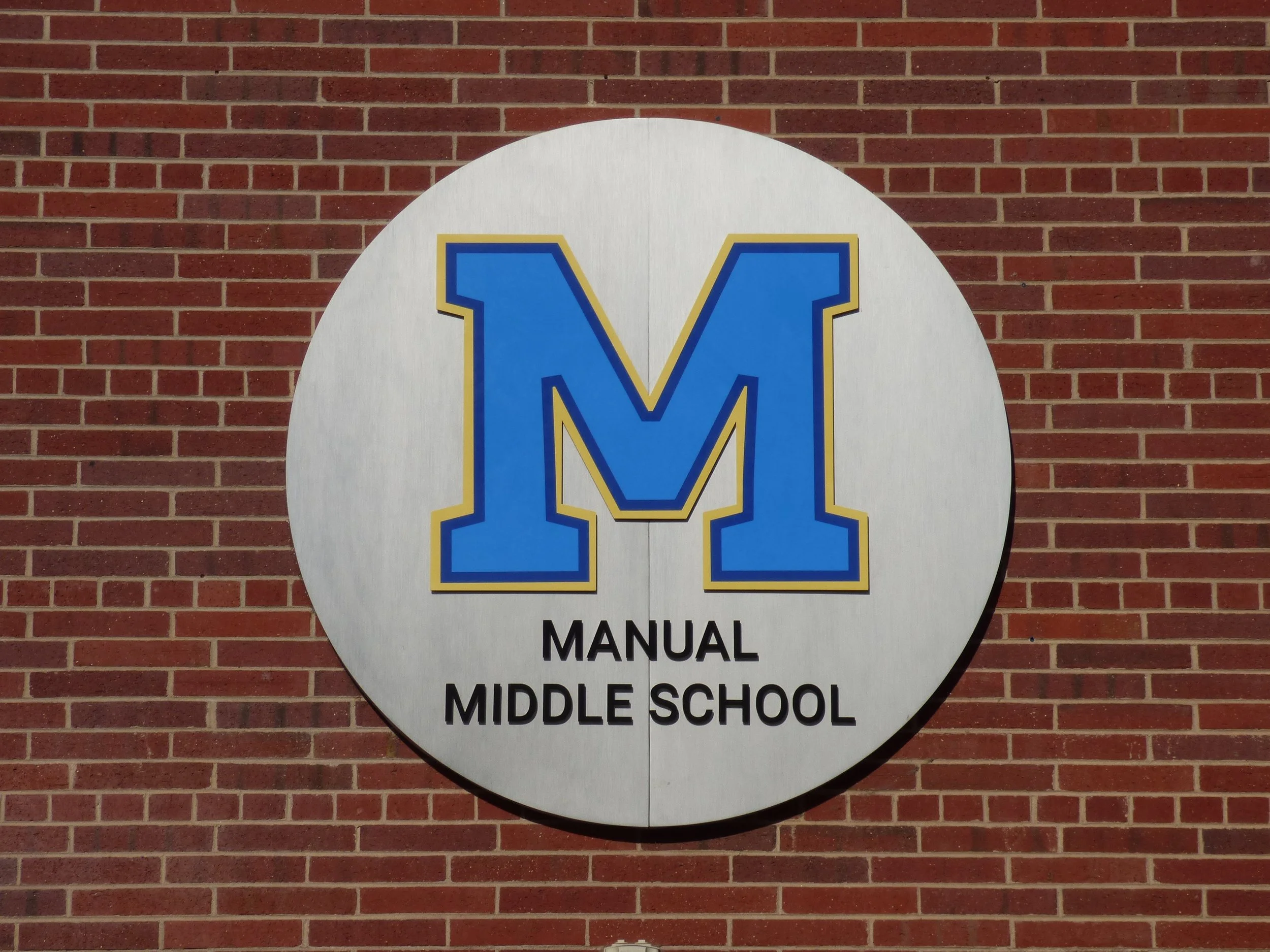 School sign with a large blue letter M outlined in gold, reading 'Manual Middle School', in front of a brick wall in Denver, CO.