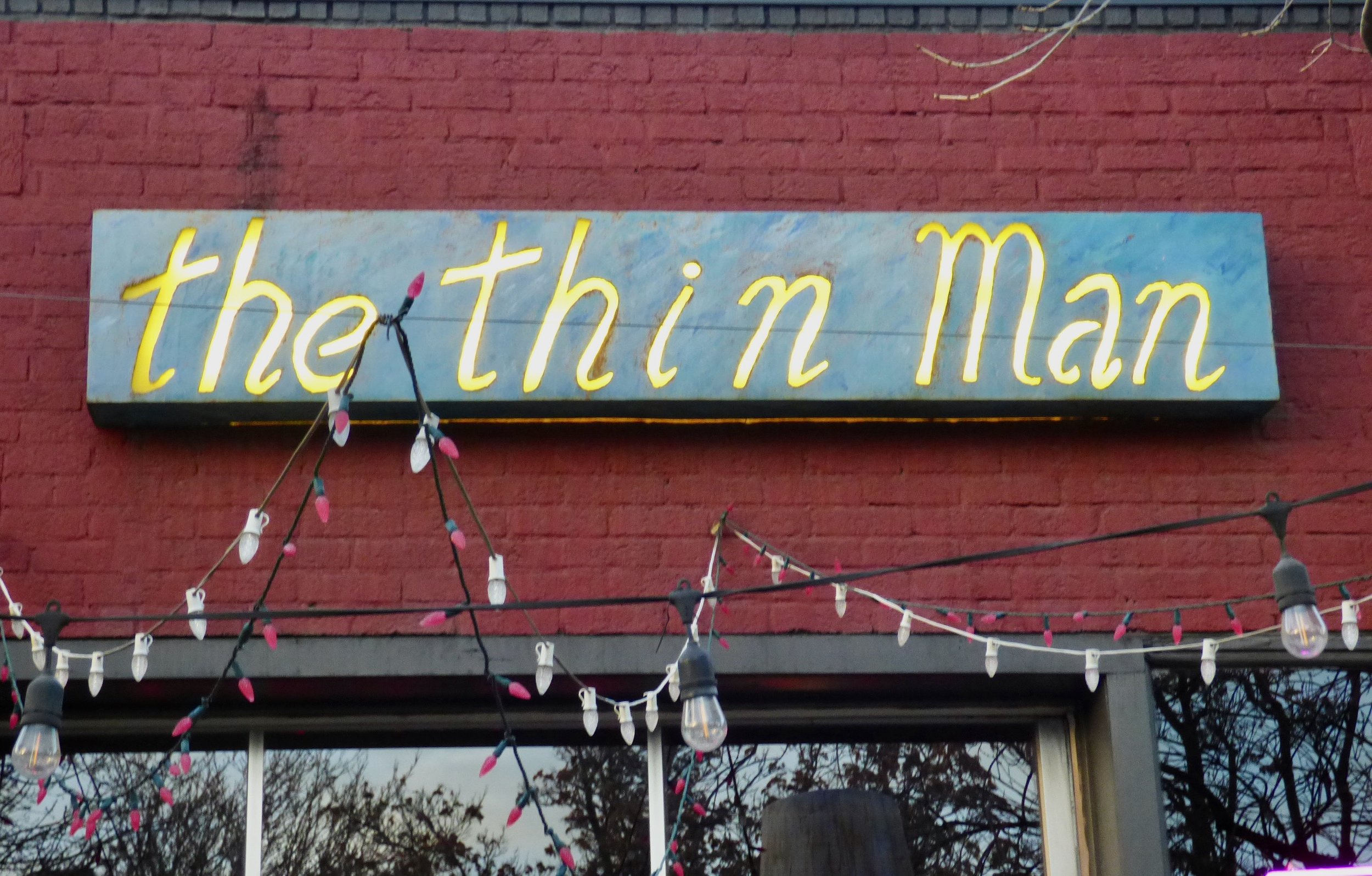 Neon sign reading 'the thin man' mounted on a red brick wall with string lights and bulbs hanging below at The Thin Man, Denver, CO.