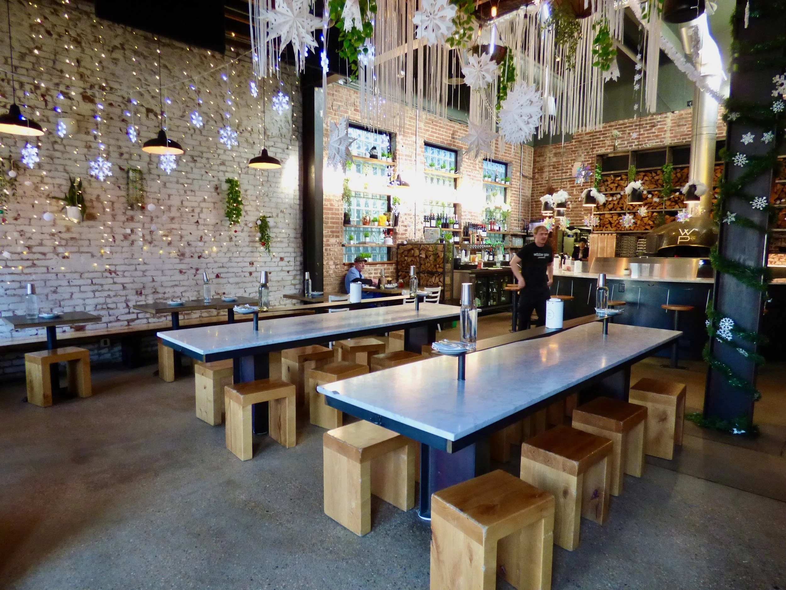 Empty restaurant interior decorated with holiday decorations including hanging snowflakes, stars, and string lights. Several tables with water bottles and napkins, and a bar area in the background.