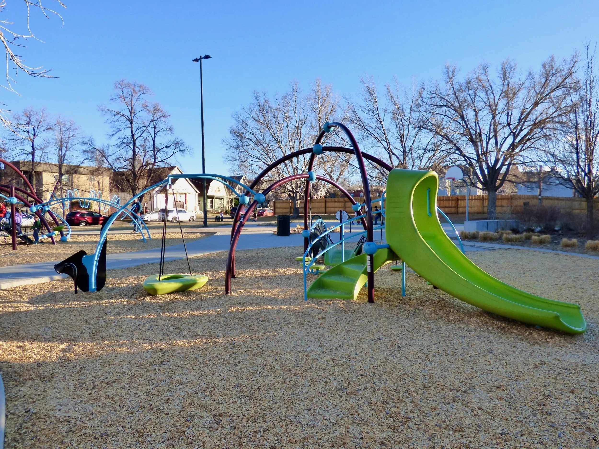 Colorful playground equipment with slides, swings, and climbing structures on a gravel surface in a park with bare trees and a clear blue sky.