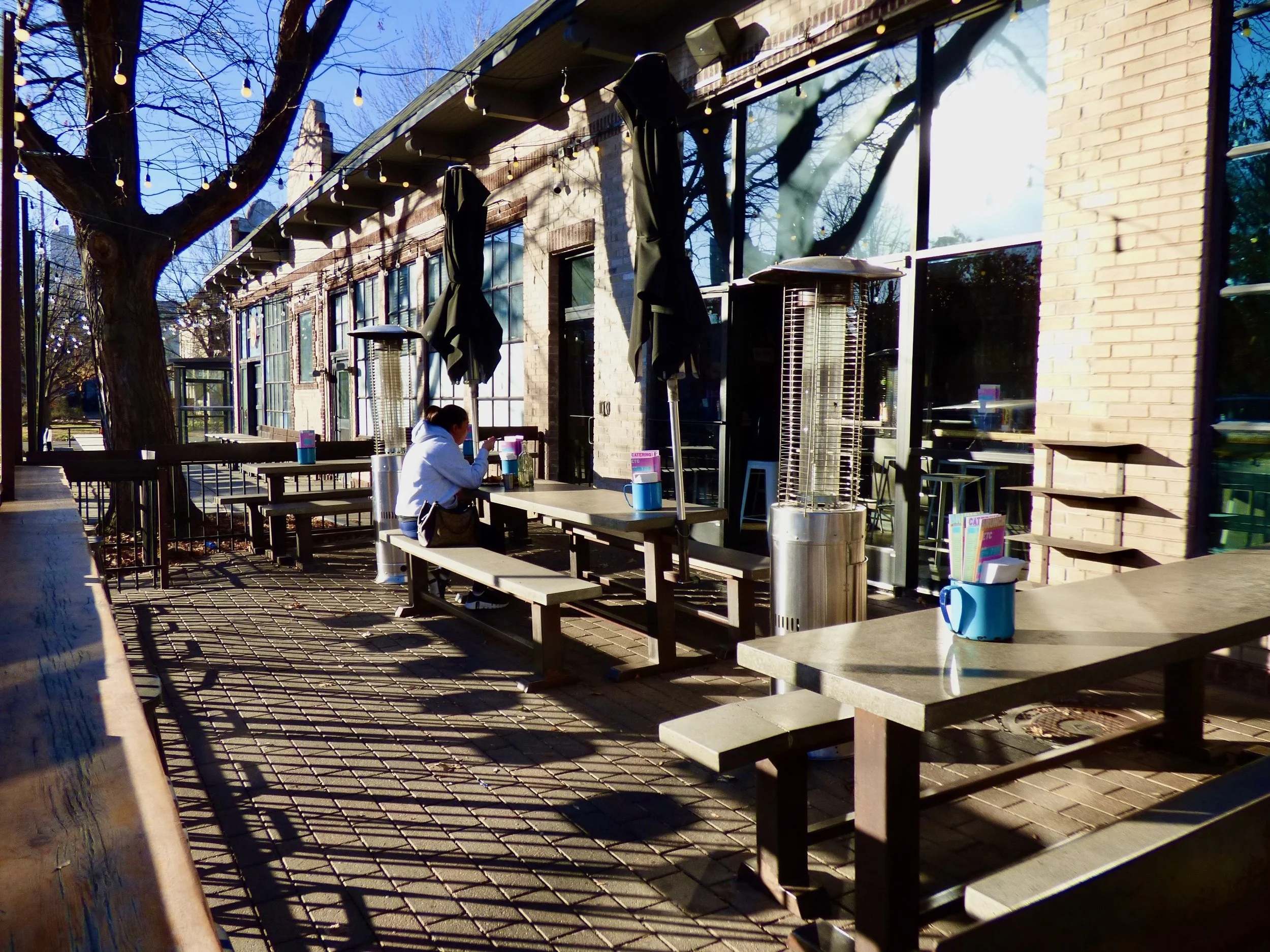 Empty outdoor patio area with wooden picnic tables, patio heaters, and a person sitting alone using a phone, with trees and a building in the background on a sunny day at Dos Santos, Denver, CO.
