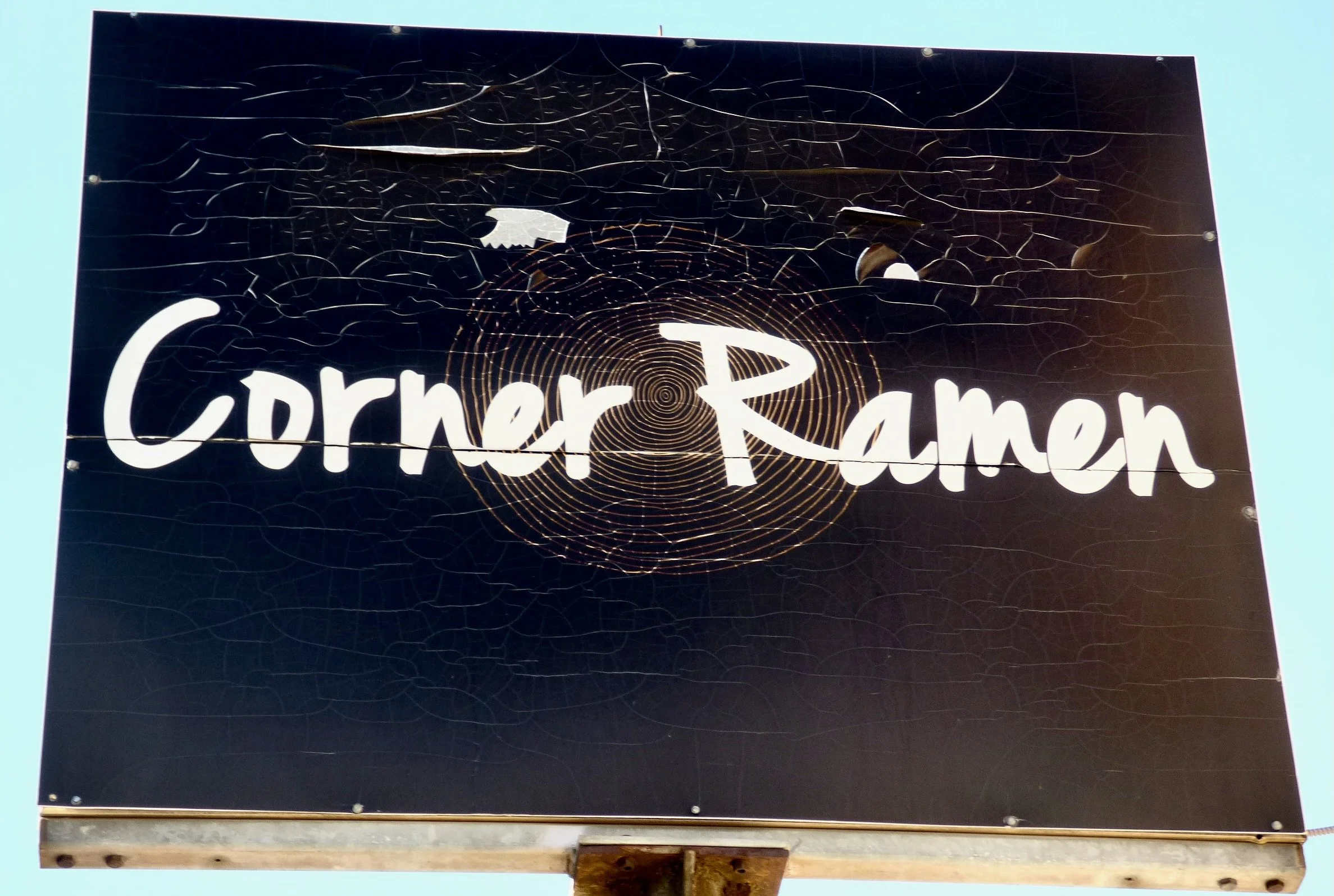 A black sign with white text that says 'Corner Ramn,' featuring a decorative spiral pattern and a web-like design, mounted on a pole against a light blue sky.