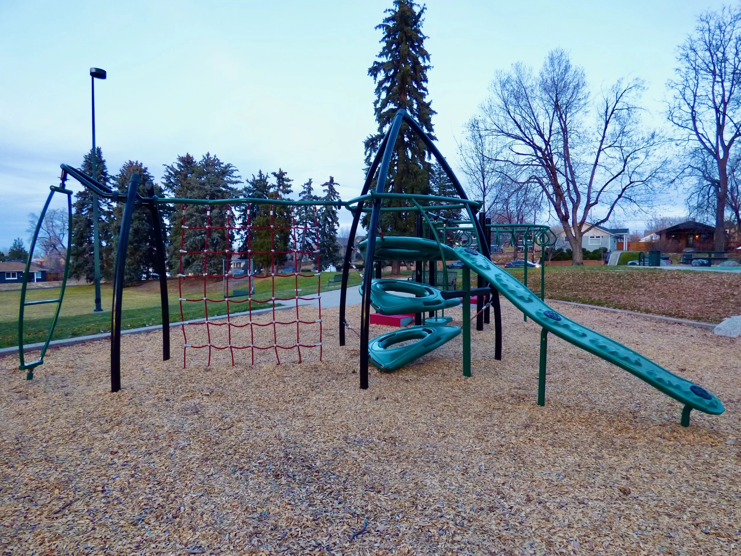 Empty playground with a green metal climbing structure, a red rope net, and a slide on a gravel surface, with leafless trees and houses in the background  in City of Nairobi Park, Denver, CO 80205.