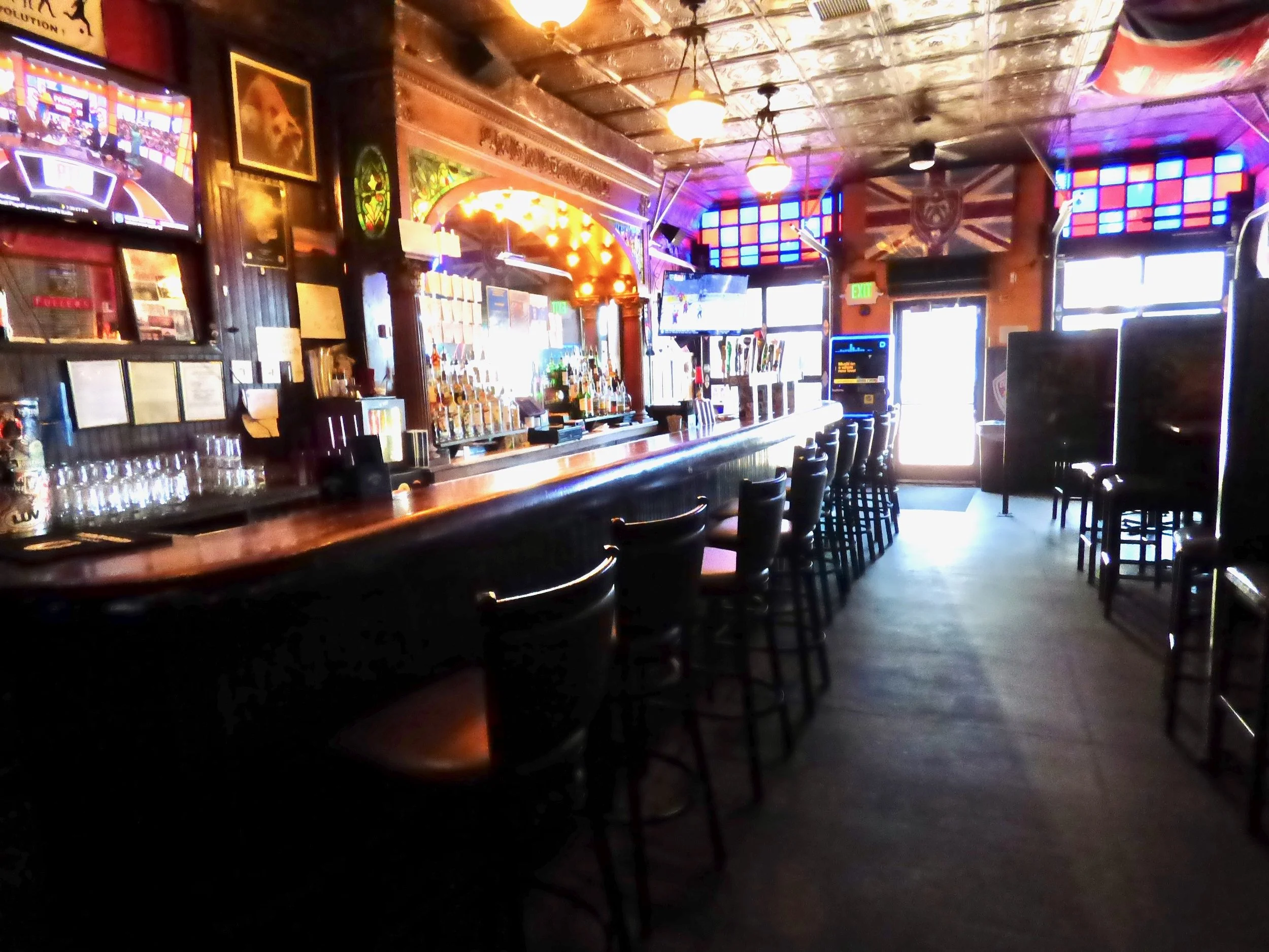 Inside a dimly lit bar with a long counter, bar stools, and various alcohol bottles behind the bar. Colorful stained glass and framed pictures adorn the walls, with multiple TV screens at The British Bulldog, Denver, CO 80205.