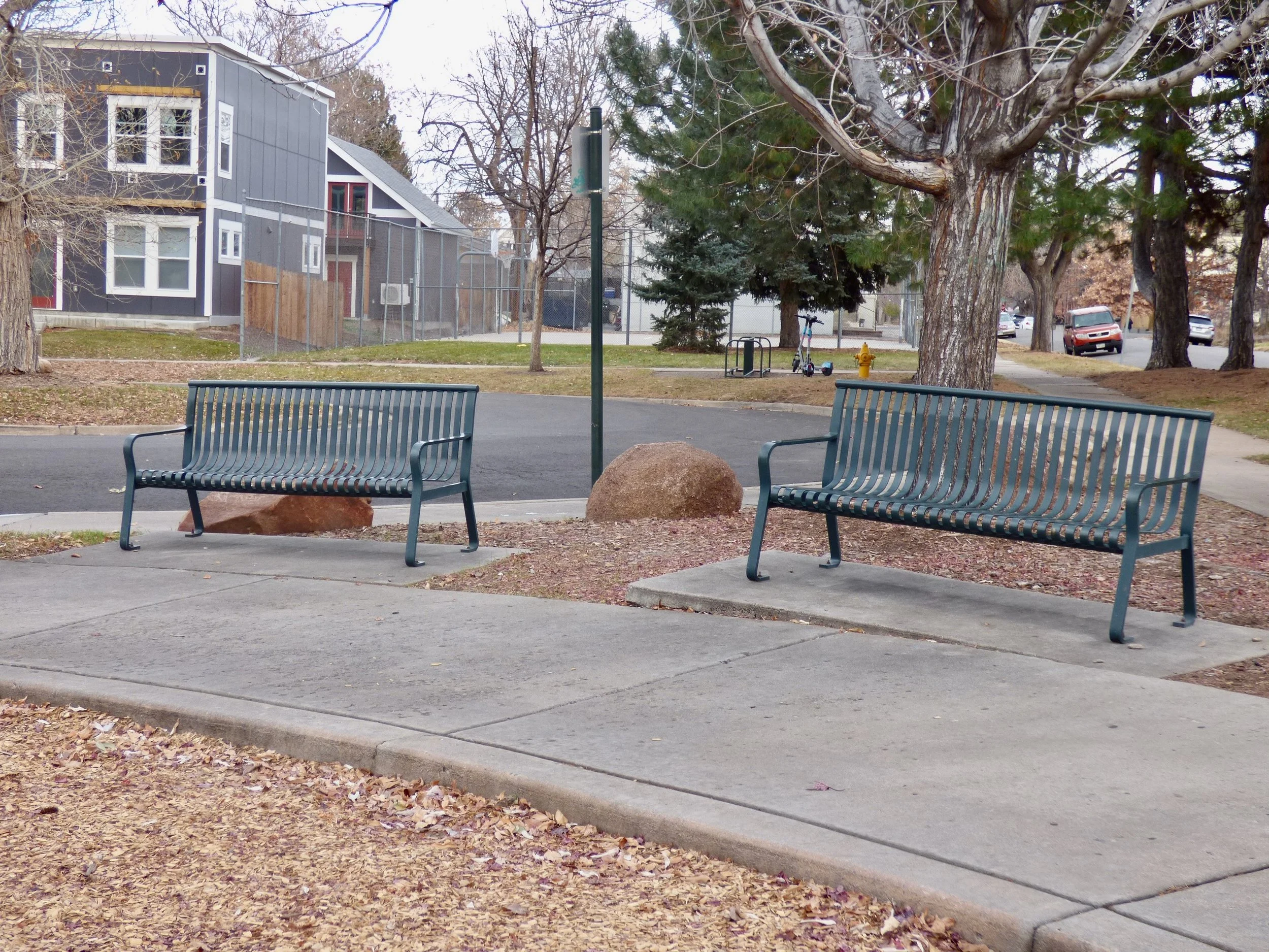 Two empty green metal benches facing each other on a sidewalk in a park, with fallen leaves on the ground and a large tree behind them at Dr. Daniel Hale Williams Park, Denver, CO 80205.