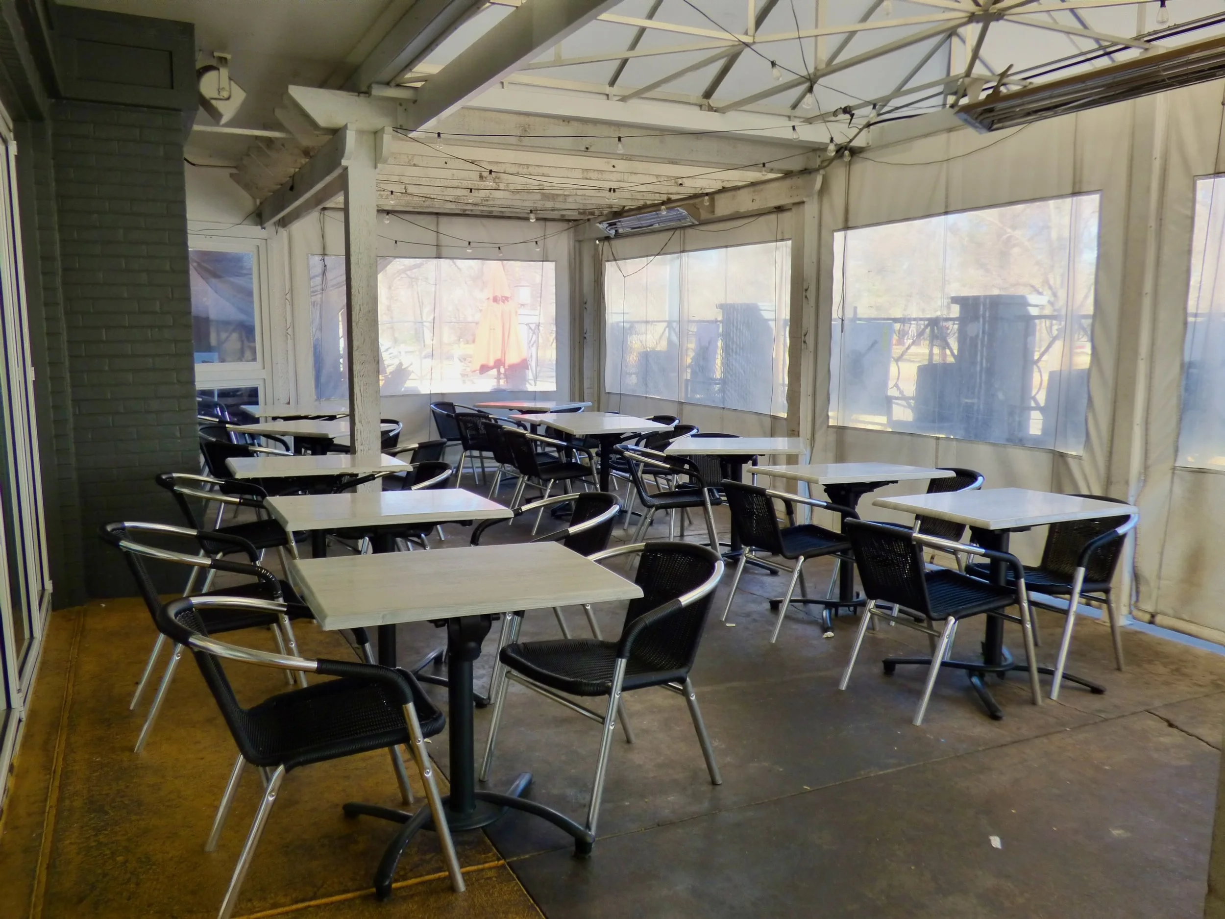 Empty outdoor seating area with white tables and black chairs inside a screened porch with large windows and string lights.