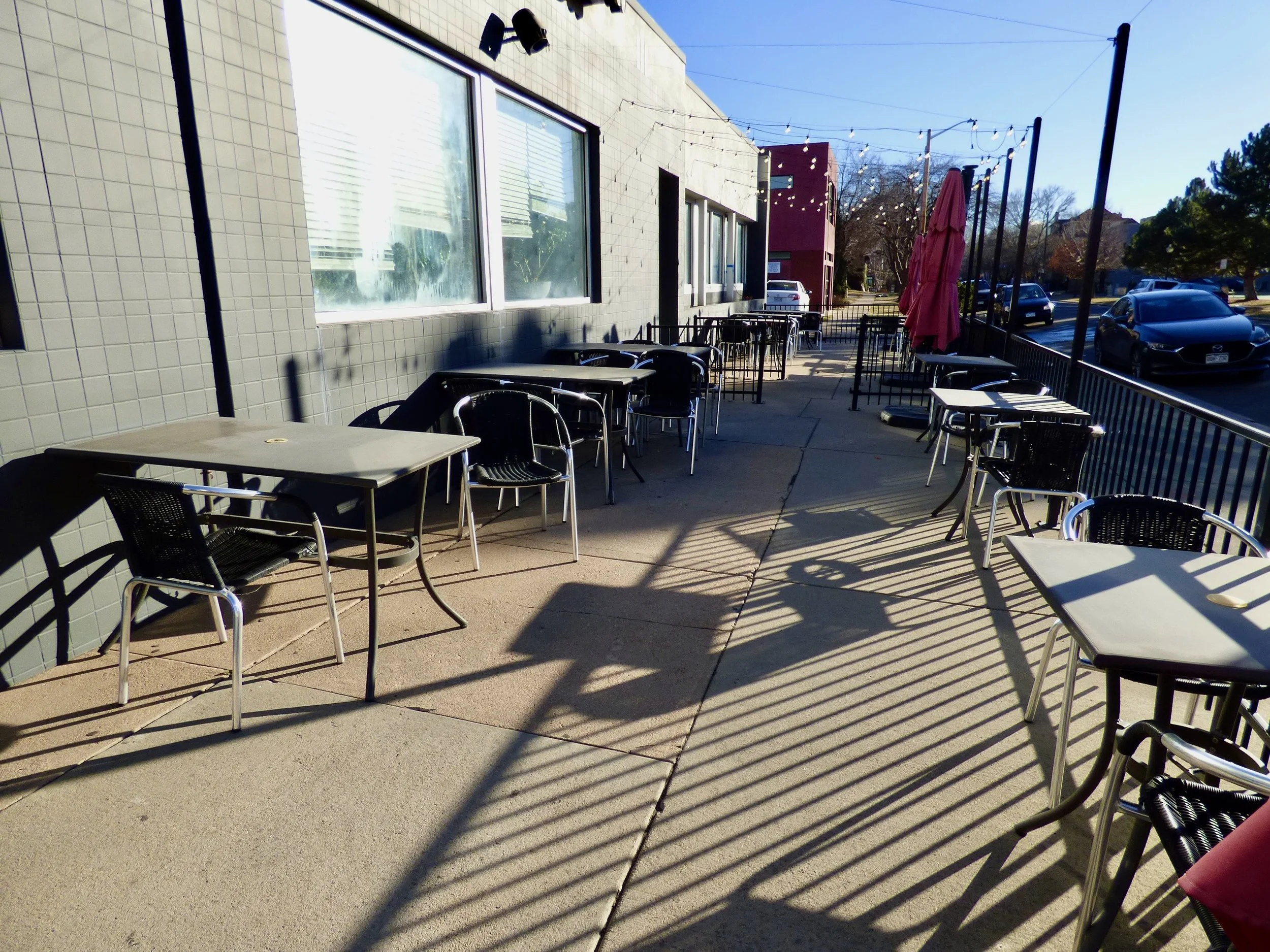 Empty outdoor patio with metal tables and chairs, sun casting shadows, string lights overhead, and closed pink umbrella.