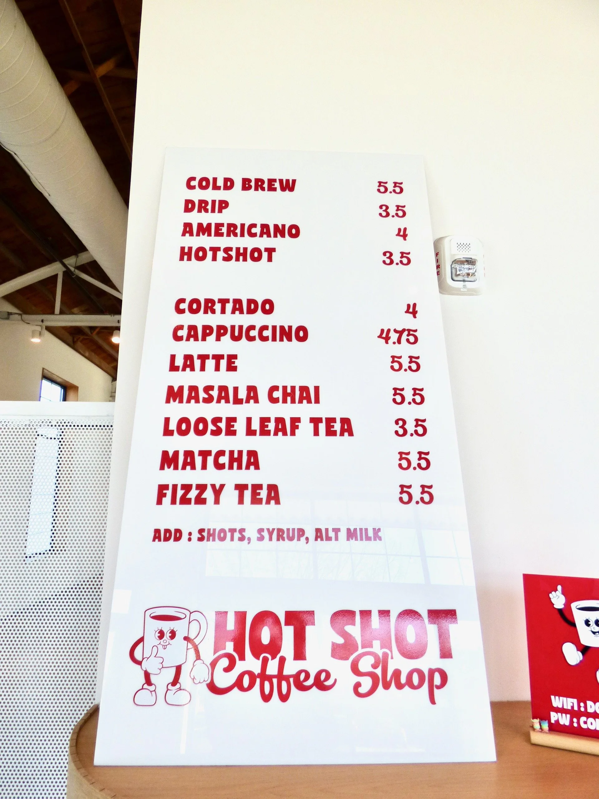 Coffee shop menu board listing drinks and prices, including cold brew, drip, americano, hotshot, cortado, cappuccino, latte, masala chai, loose leaf tea, matcha, and fizzy tea, with an option to add shots at Hot Shot Coffee Shop, Denver, CO 80205.