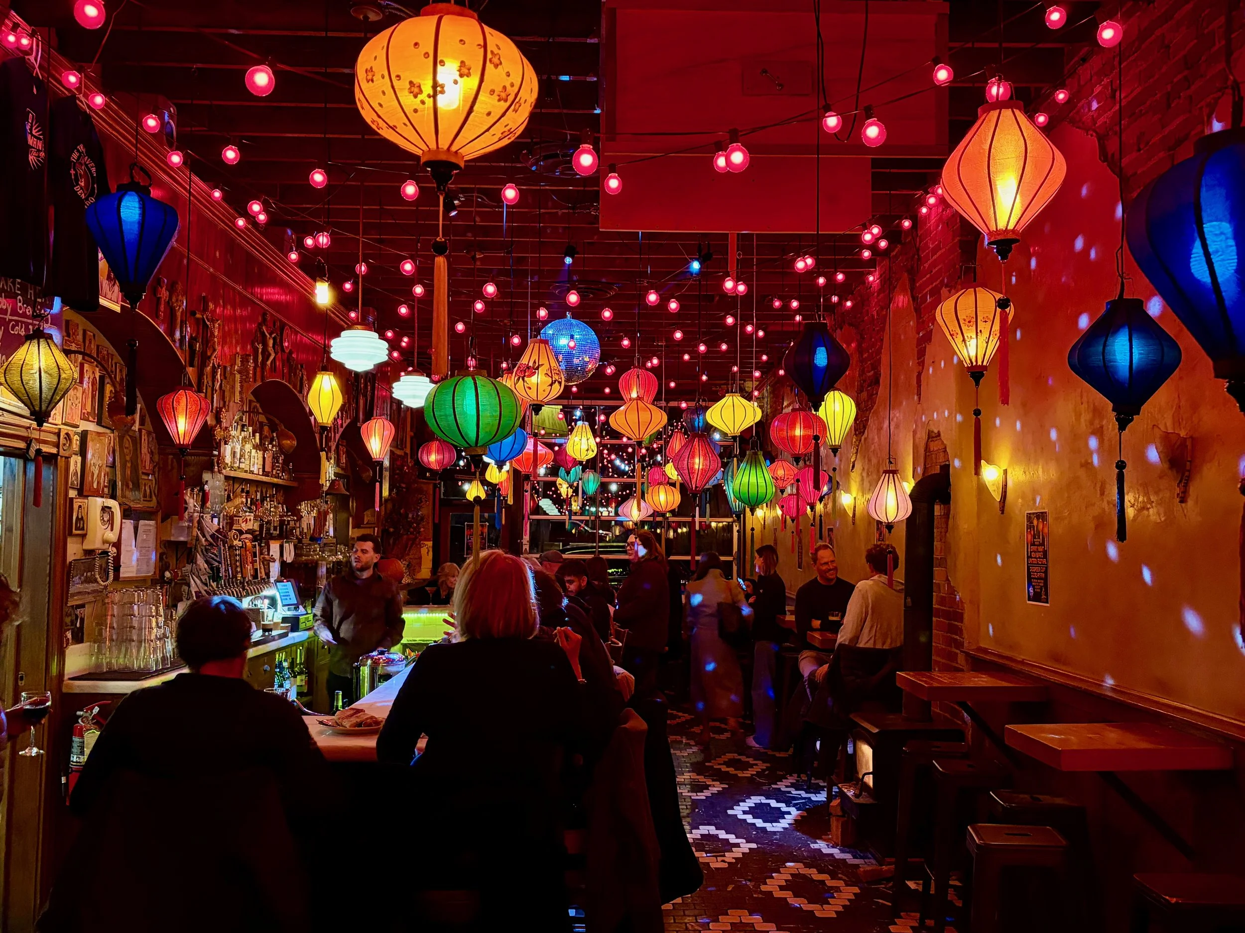 Inside a colorful bar decorated with multicolored hanging lanterns and string lights, with several patrons sitting and standing, and a bartender behind the bar at The Thin Man, Denver, CO.