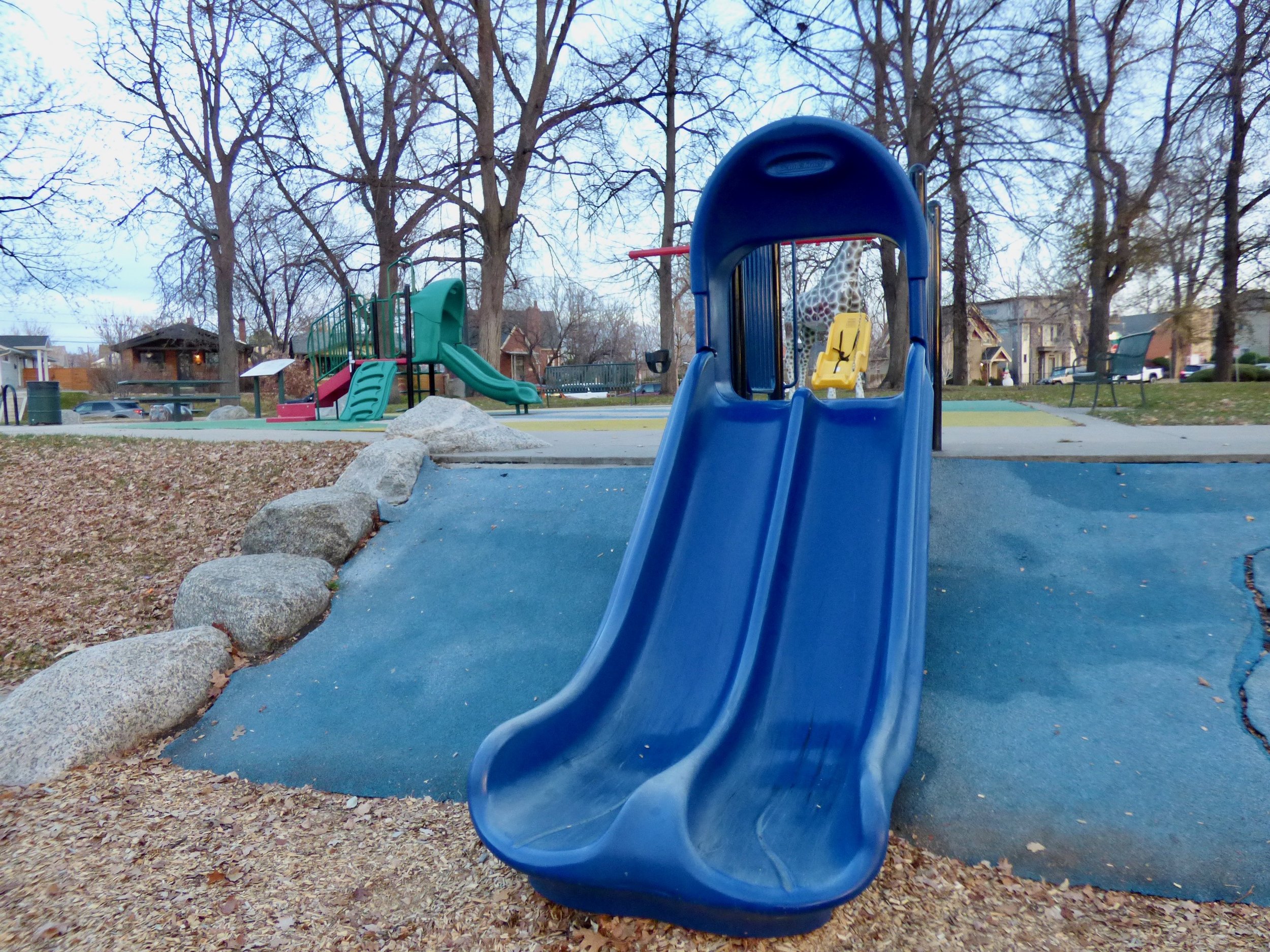Colorful playground with slides and climbing structures in an outdoor park, with trees and houses in the background in City of Nairobi Park, Denver, CO 80205.