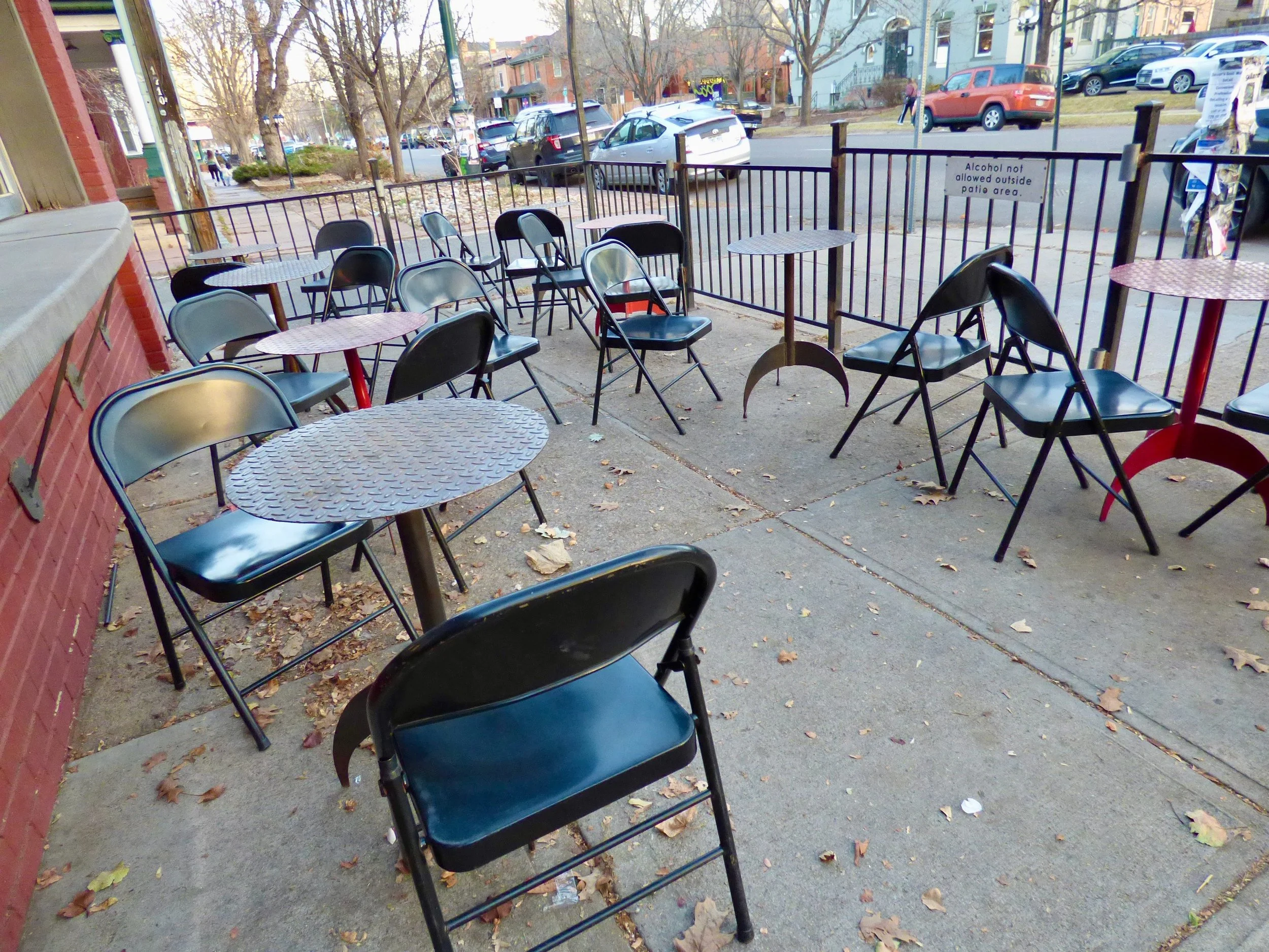 Empty outdoor patio with metal tables and chairs, some tipped over, surrounded by a black metal fence with a sign restricting alcohol outside at St. Mark's Coffeehouse and Bakery, Denver, CO.