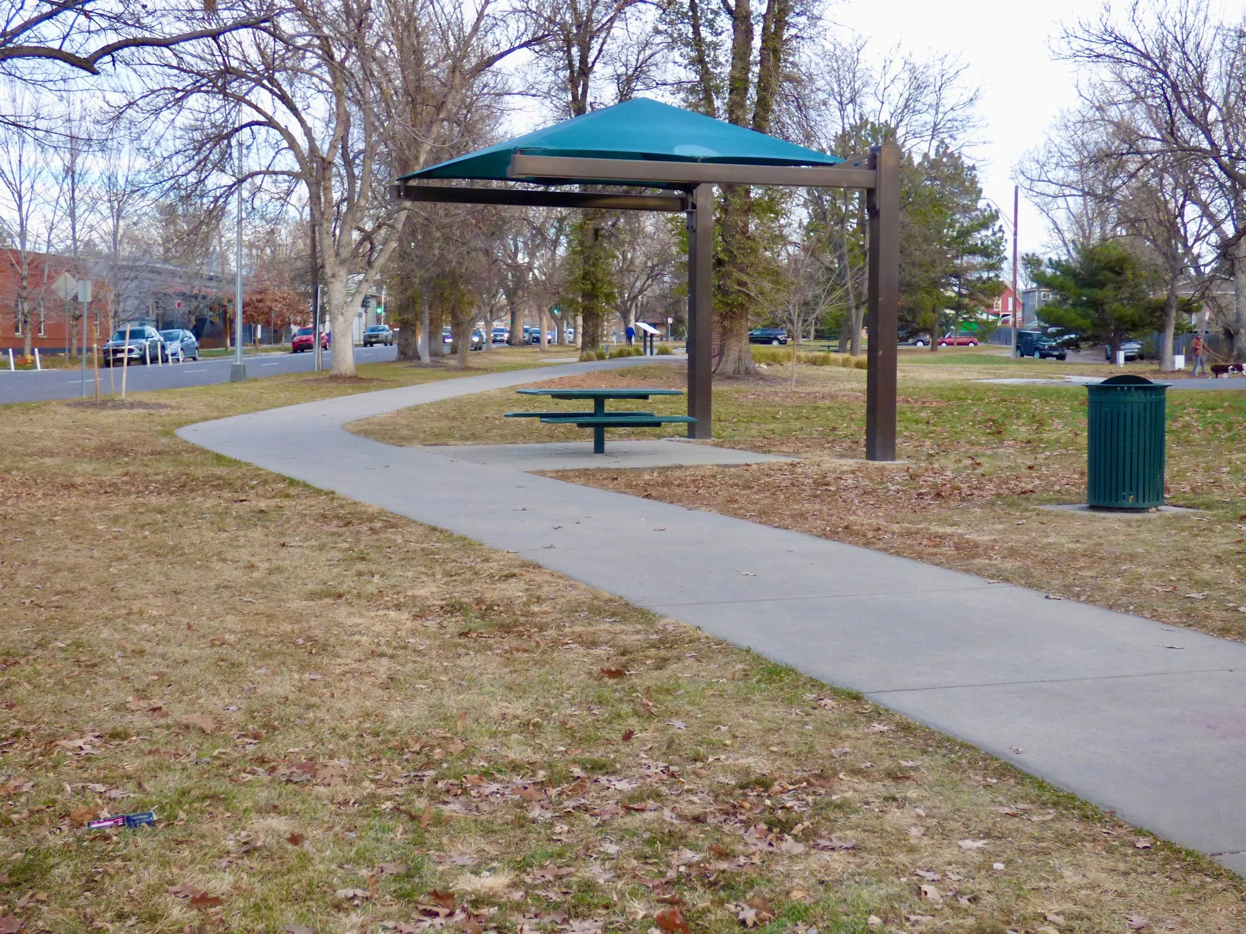 A park with a sidewalk, a picnic table under a shade structure, a trash can, leafless trees, and cars on the street in the background at George Morrison Sr. Park, Denver, CO 80205.
