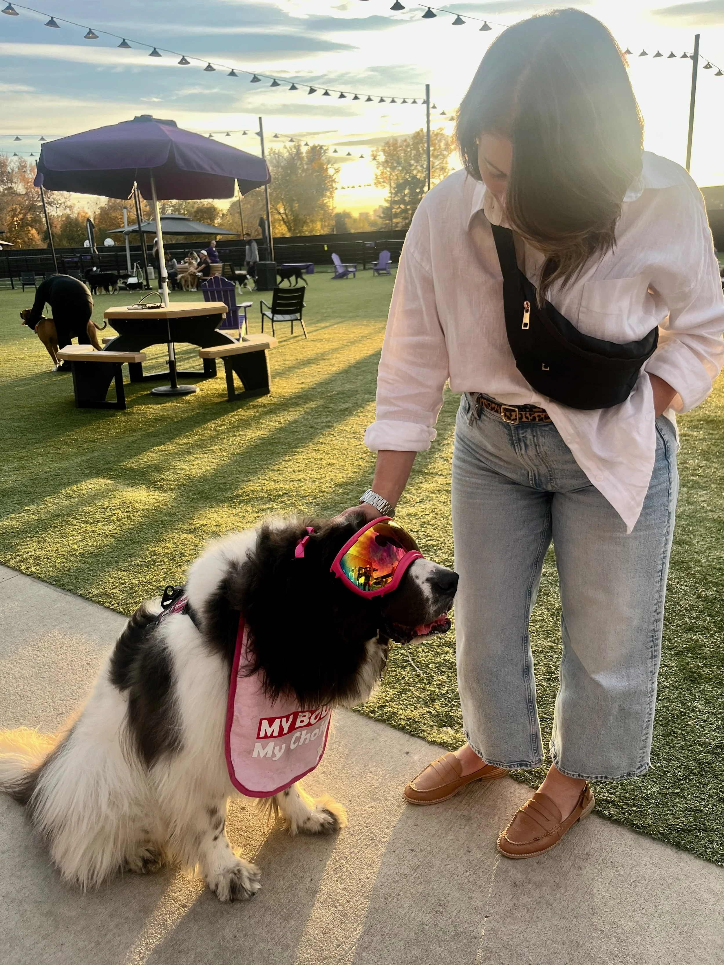 A woman with short brown hair wearing a white shirt, light blue jeans, and tan loafers standing on a sidewalk with a black and white dog wearing pink sunglasses at Skiptown, Denver, CO 80205.