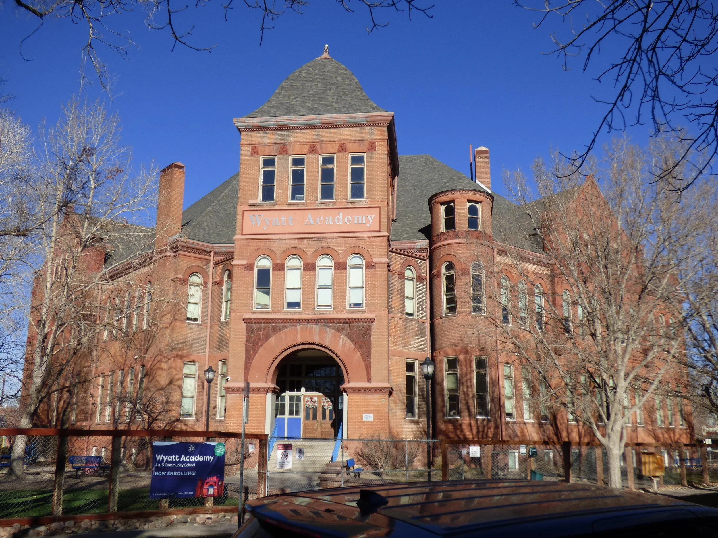 Front view of a historic red brick building with arched windows and a central entrance, labeled 'Wyatt Academy,' surrounded by leafless trees against a clear blue sky.