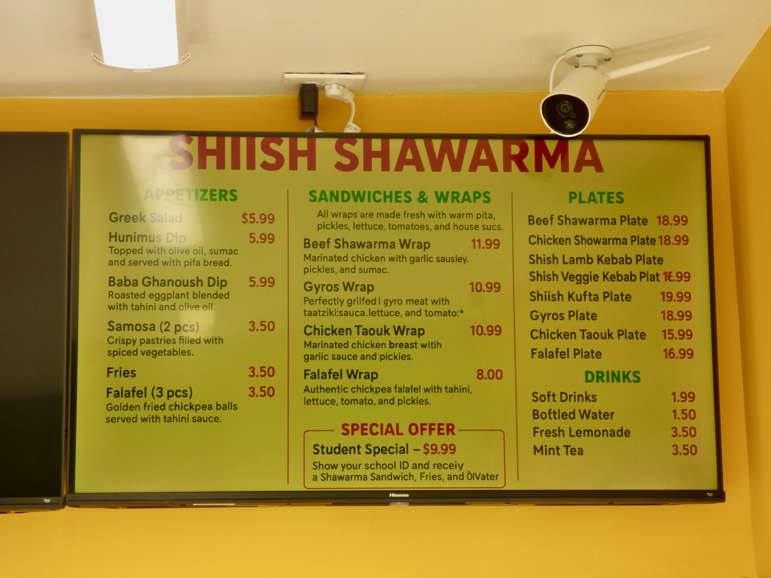 Menu board for Shish Shawarma featuring appetizers, sandwiches, plates, and drinks. Prices range from $1.50 to $19.99, with options including Greek salad, humus dip, baba ghanoush dip, samosas, fries, falafel, beef shawarma, chicken shawarma, gyros, and vegetarian plates, as well as soft drinks, bottled water, lemonade, and mint tea.