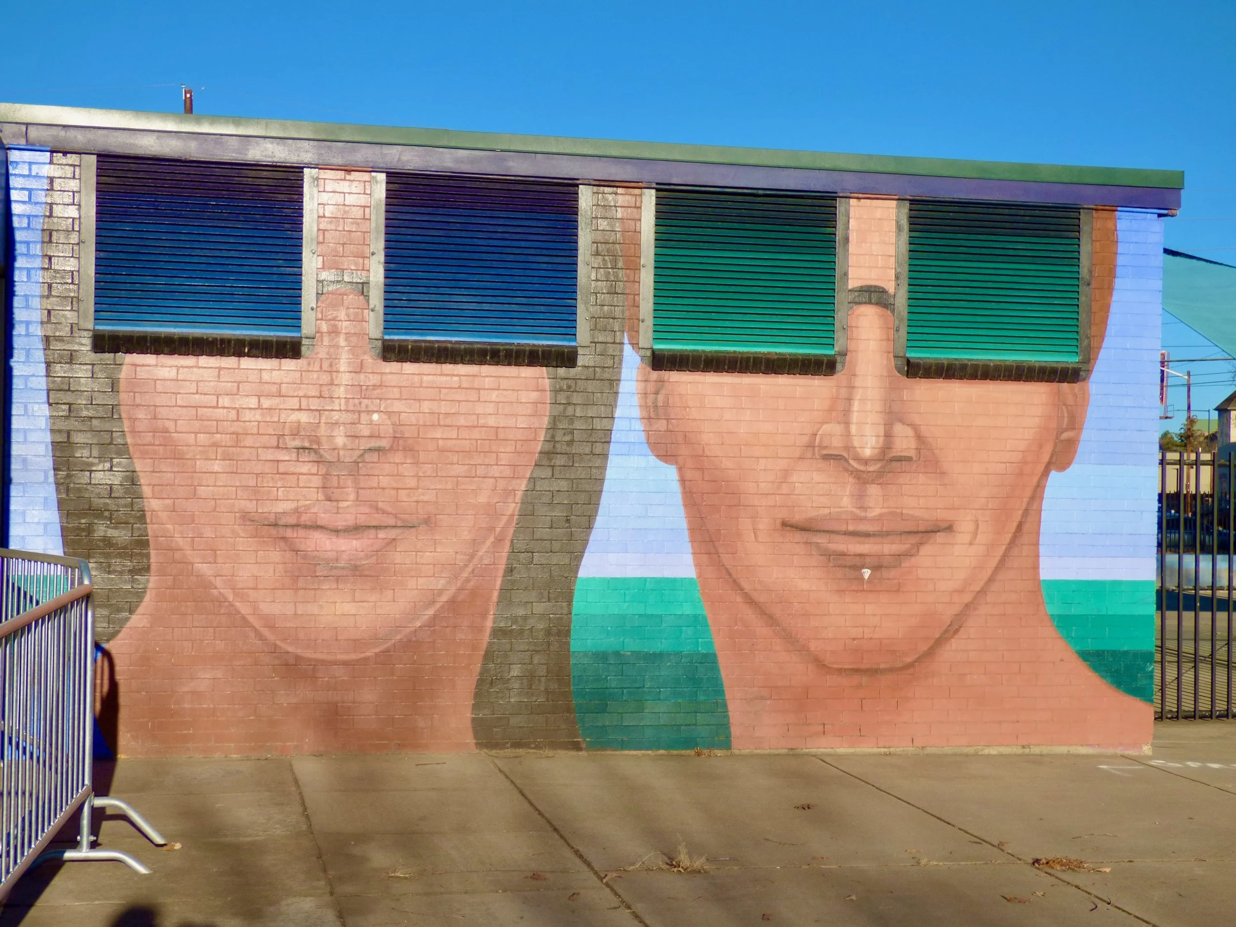 Mural of two women's faces on a brick wall, each wearing colorful sunglasses with a blue sky background.