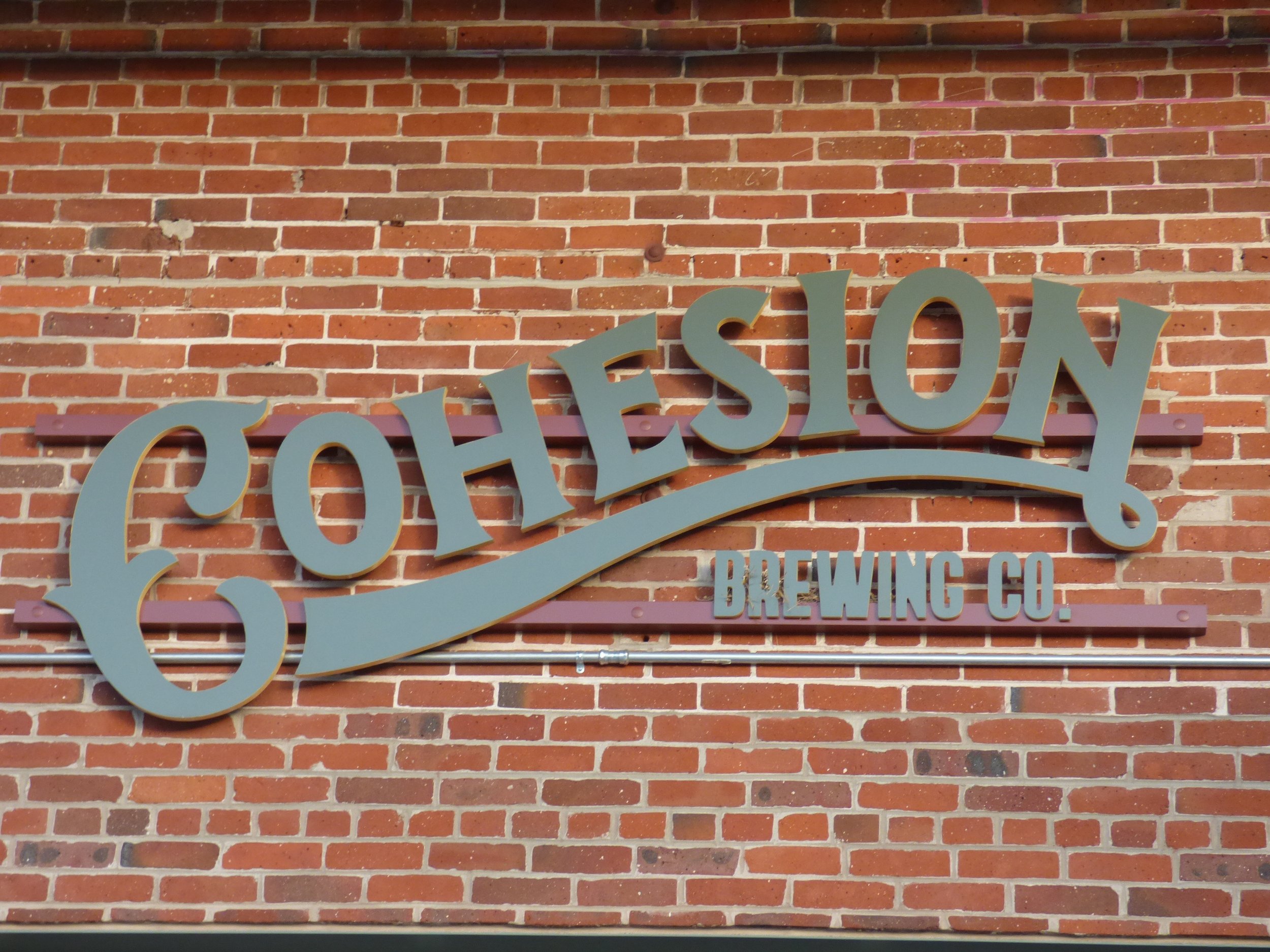 Sign for Cohesion Brewing Co. mounted on a red brick wall with beige and brown bricks at Cohesion Brewery, Denver, CO 80205.