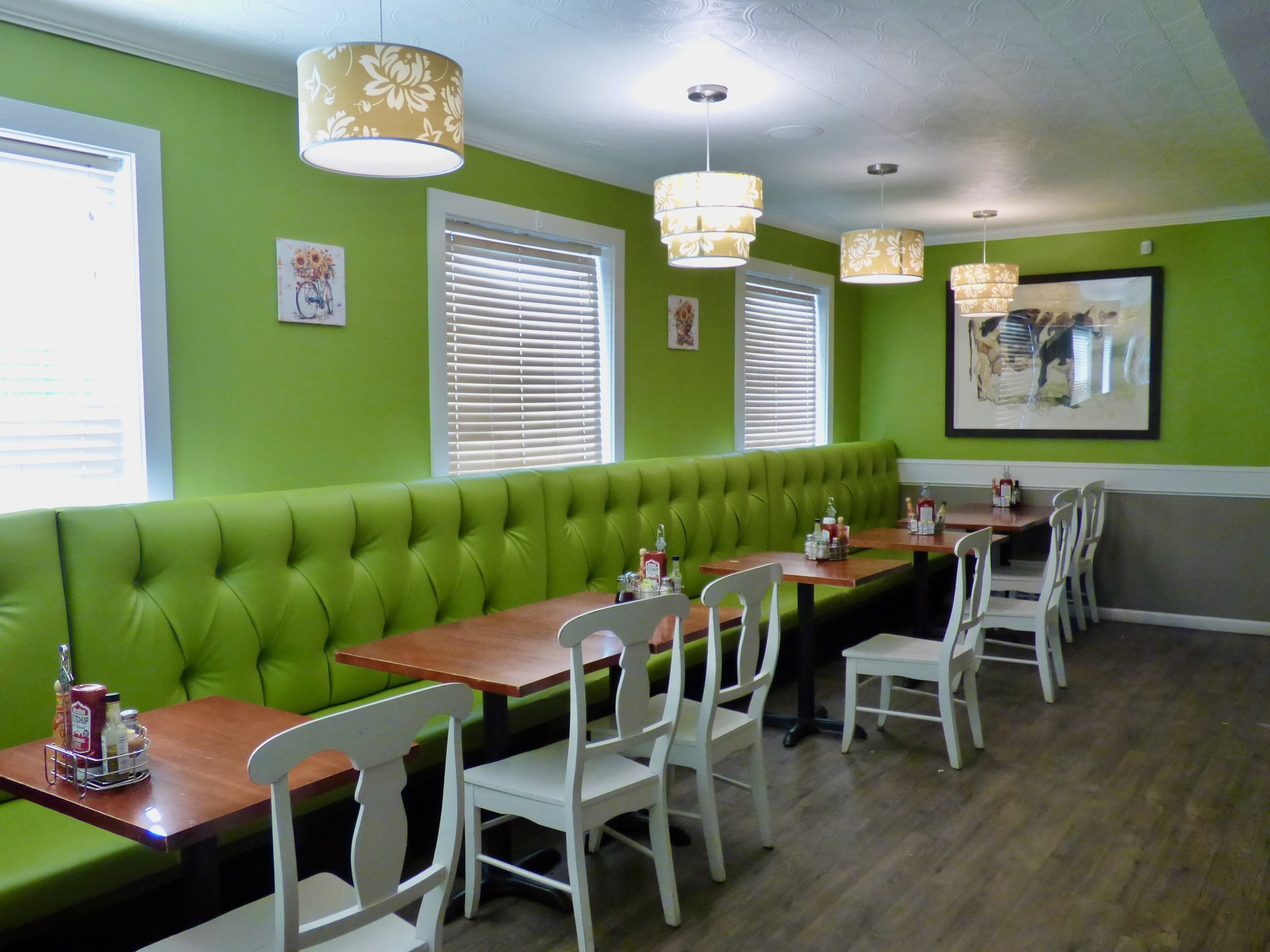 Interior of a restaurant with a long green cushioned bench along the wall, white chairs at wooden tables, and four windows with white blinds. There are framed pictures on the lime green wall and ceiling lights with patterned shades.