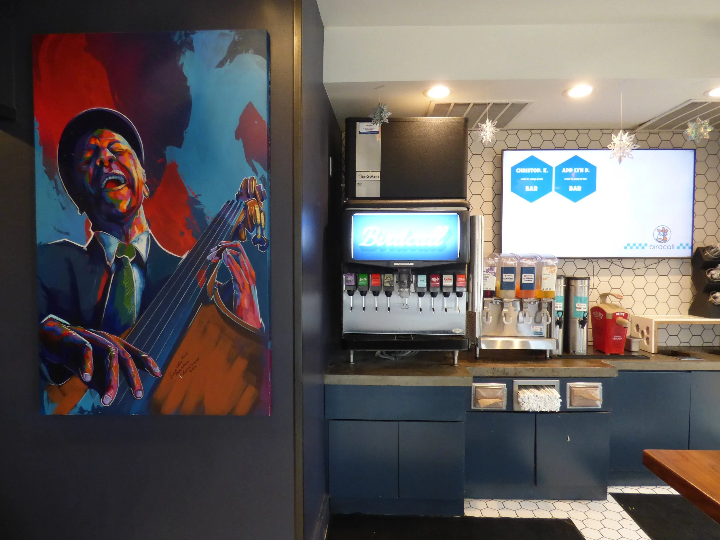 Colorful mural of a man playing guitar in a jazz style, located in a café or restaurant with beverage dispensers and a digital menu screen.