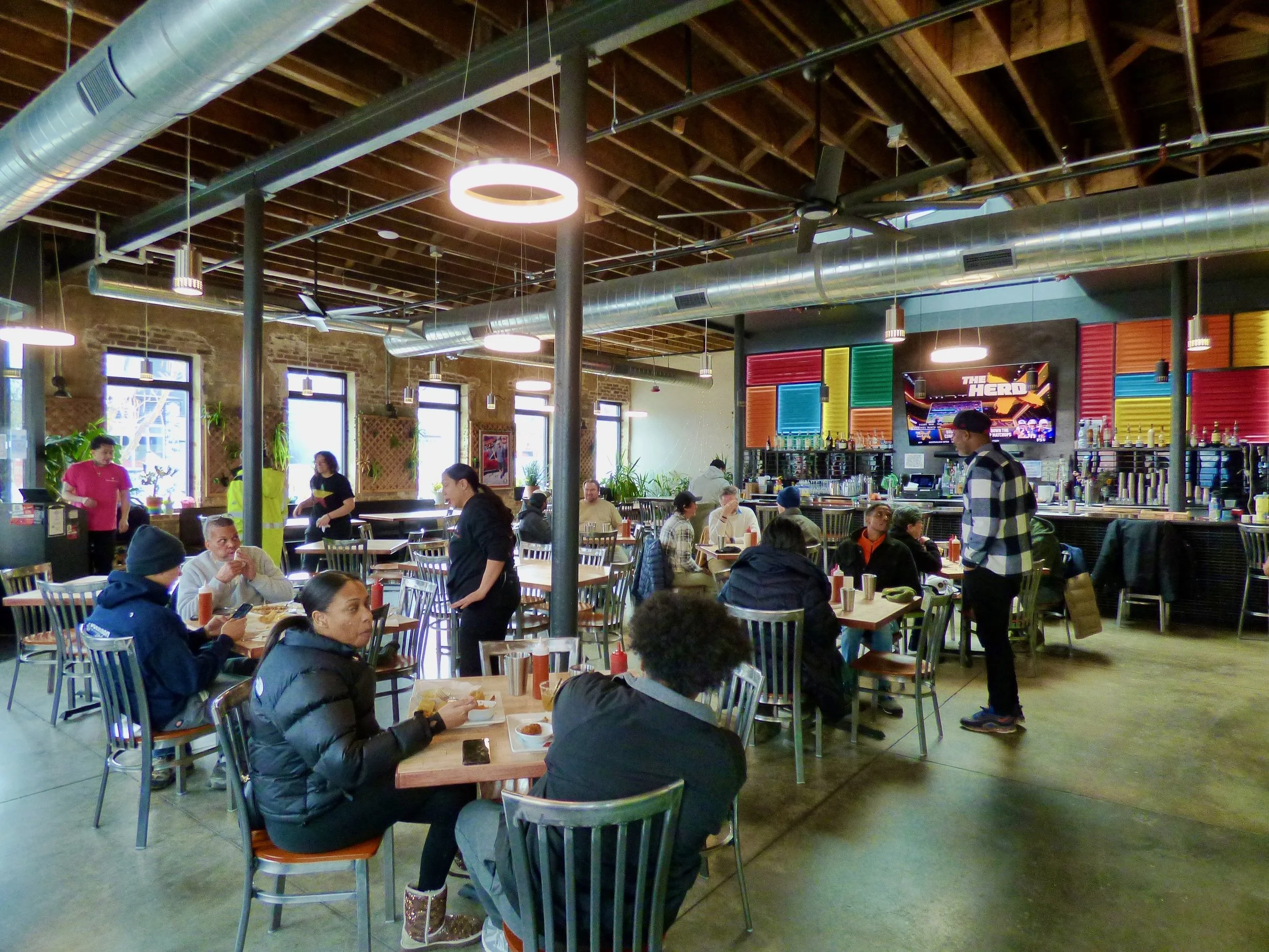 Inside a busy restaurant with several customers seated at tables, some are eating and others are chatting, with wait staff attending to them. The restaurant has high ceilings with exposed wooden beams and large industrial-style metal ducts, colorful wall decor behind the bar, and large windows letting in natural light.