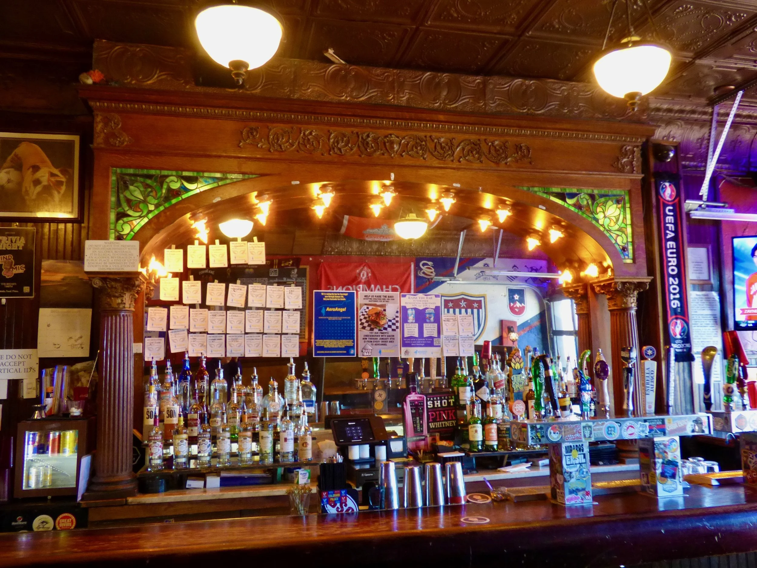 A bar with a variety of alcohol bottles, a cash register, and promotional signs, with a large mirror and wood-paneled decor at The British Bulldog, Denver, CO 80205.