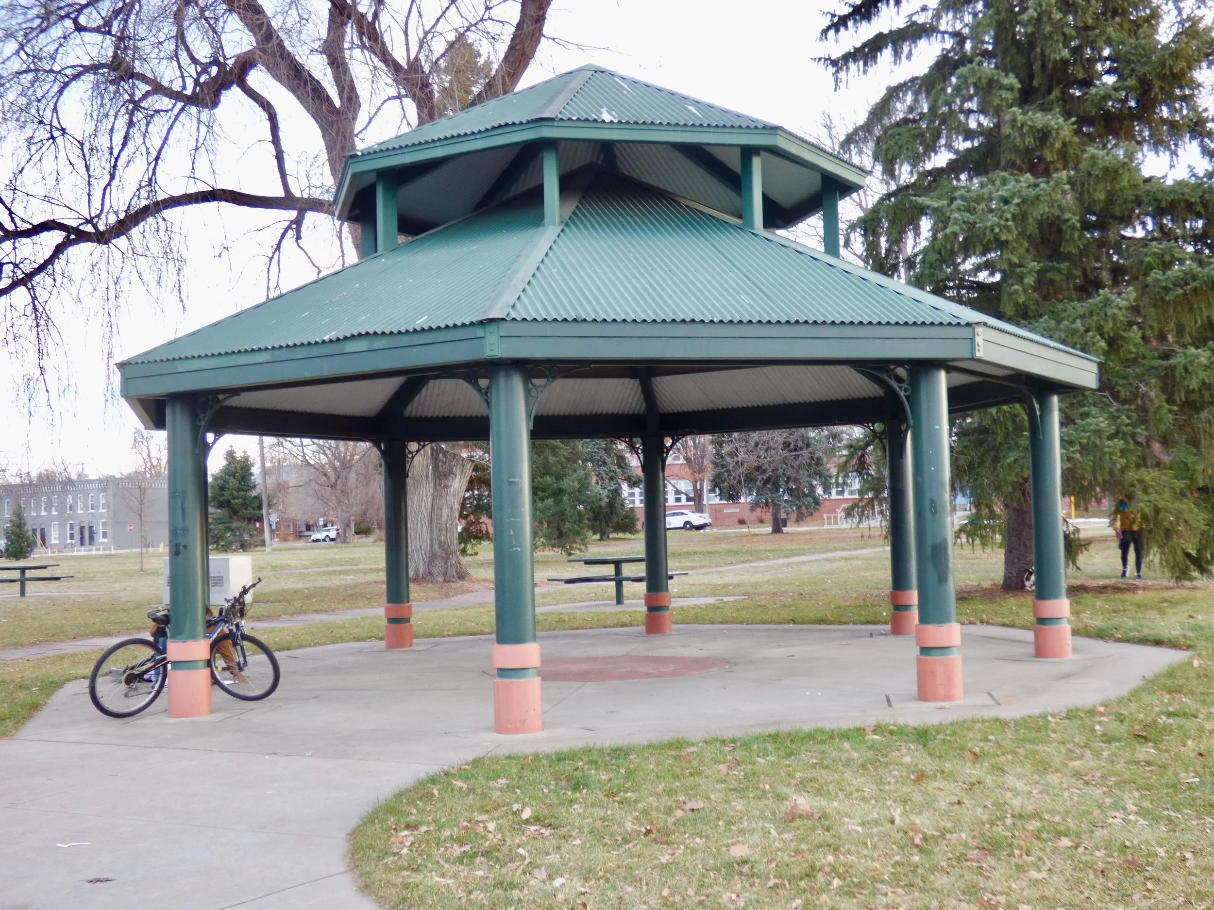 A green park pavilion with a tin roof, supported by columns, located in a grassy park. There is a bicycle parked next to the pavilion. Trees and a few people are visible in the background at Fuller Park, Denver, CO 80205.