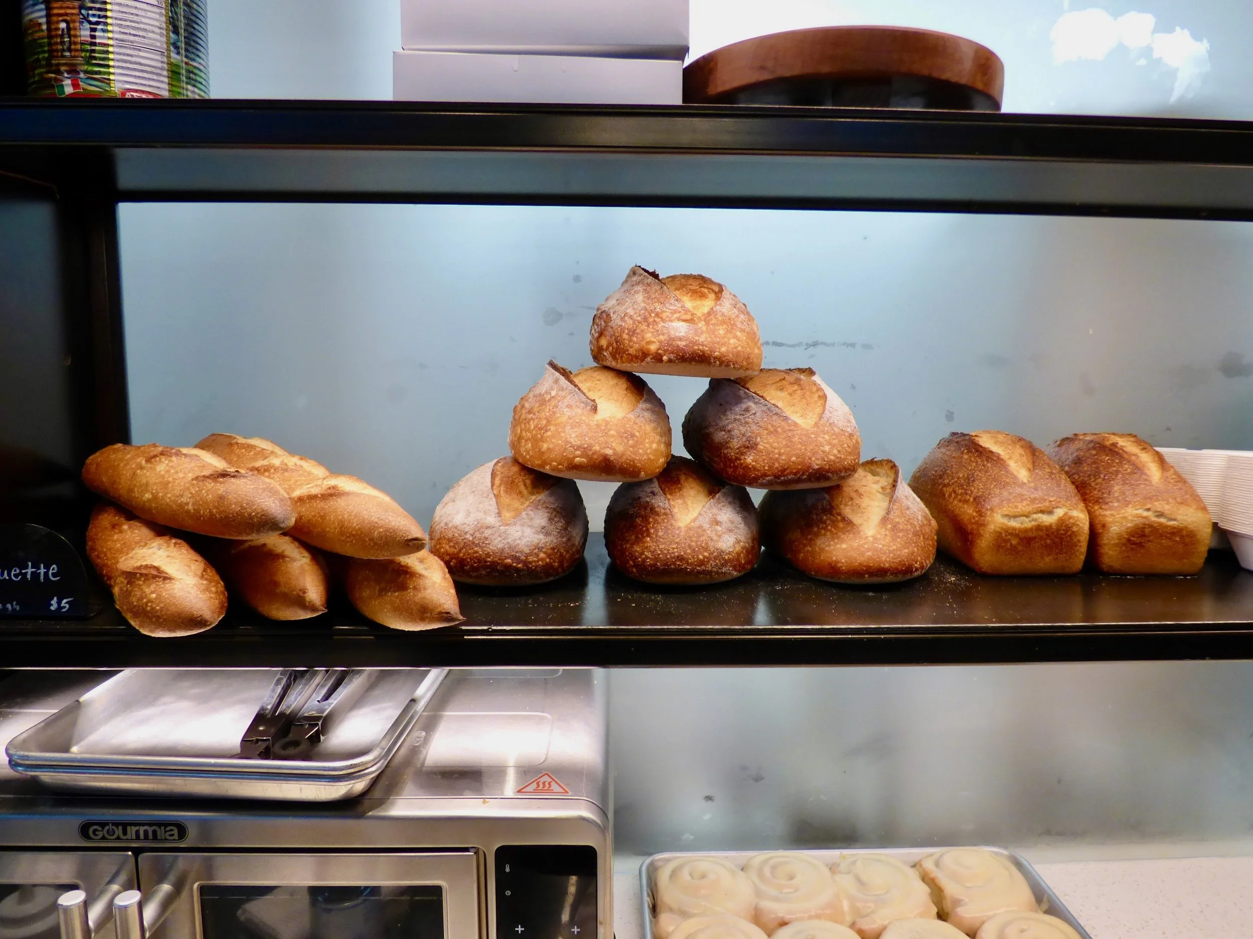 Various types of fresh bread and baked goods displayed on a glass shelf in a bakery or cafe, including baguettes and round bread loaves at Elemental Bakery & Coffeehouse, Denver, CO 80205.
