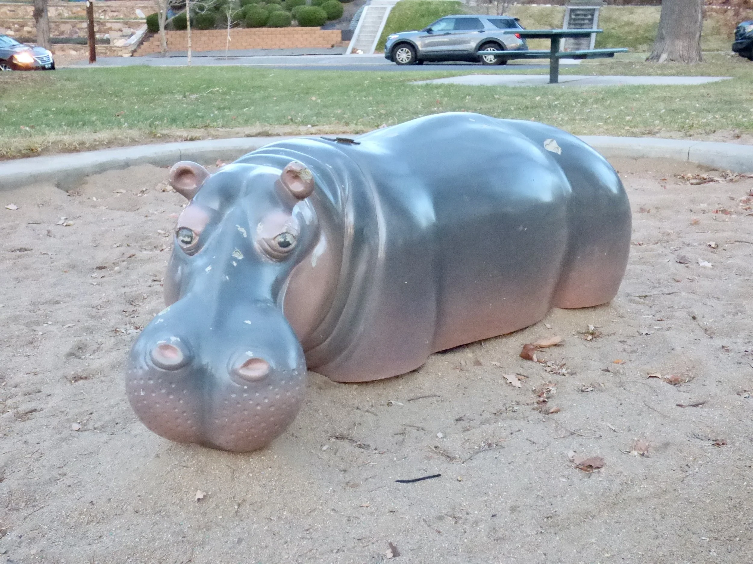 A metal sculpture of a hippopotamus lying on a sandy ground in a park.