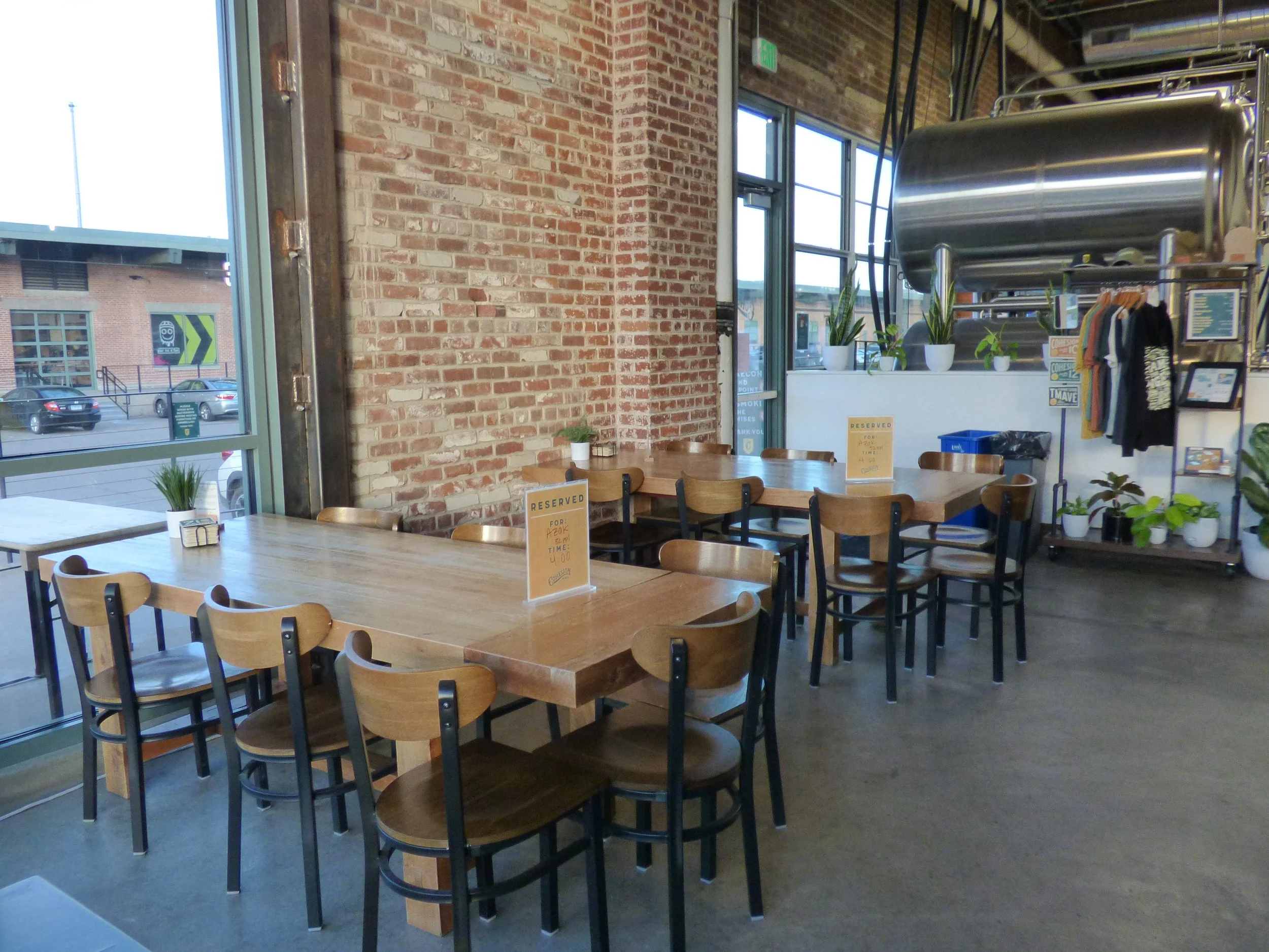 Empty restaurant with wooden tables, chairs, and potted plants, brick walls, large windows, and a metal kitchen apparatus in the background at Cohesion Brewery, Denver, CO 80205.