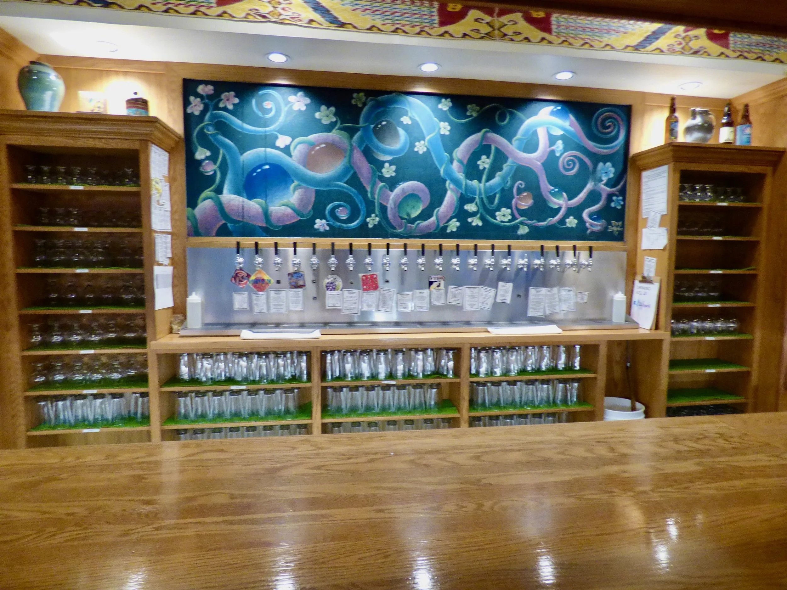 A taproom with multiple taps and shelves of glasses, against a mural of twisting vines and flowers in blue and purple hues at Vine Street Pub, Denver, CO.